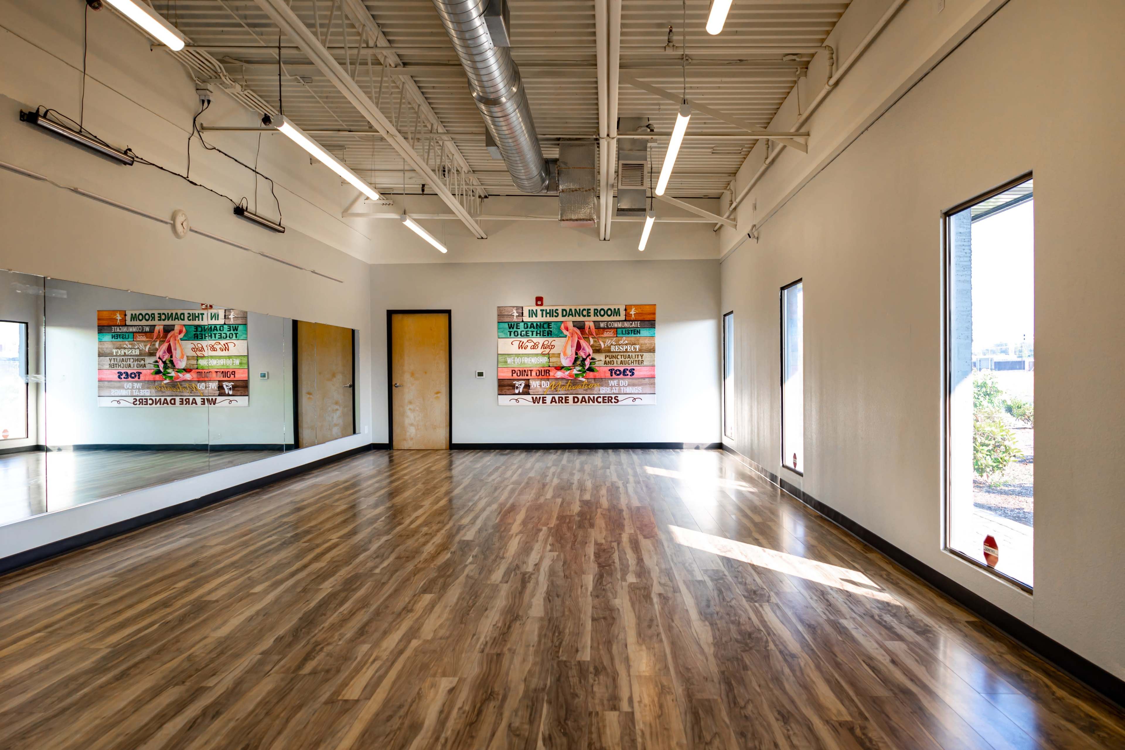 A spacious dance studio with a large mirror on one wall and two windows providing natural light.
