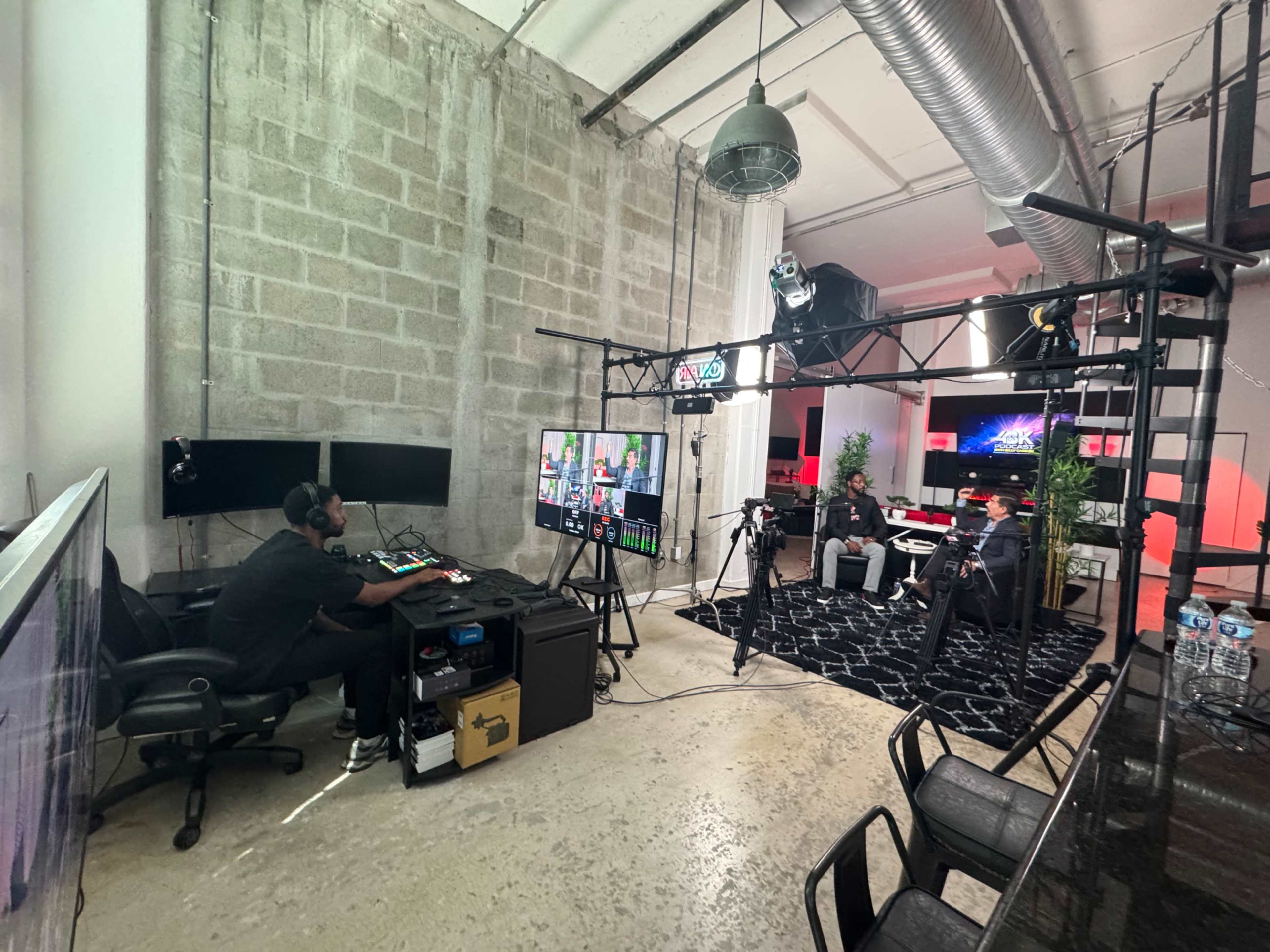 The image shows a modern studio setting with a crew operating video equipment while two people are seated for an interview or discussion on a set designed with lighting and decorative elements.