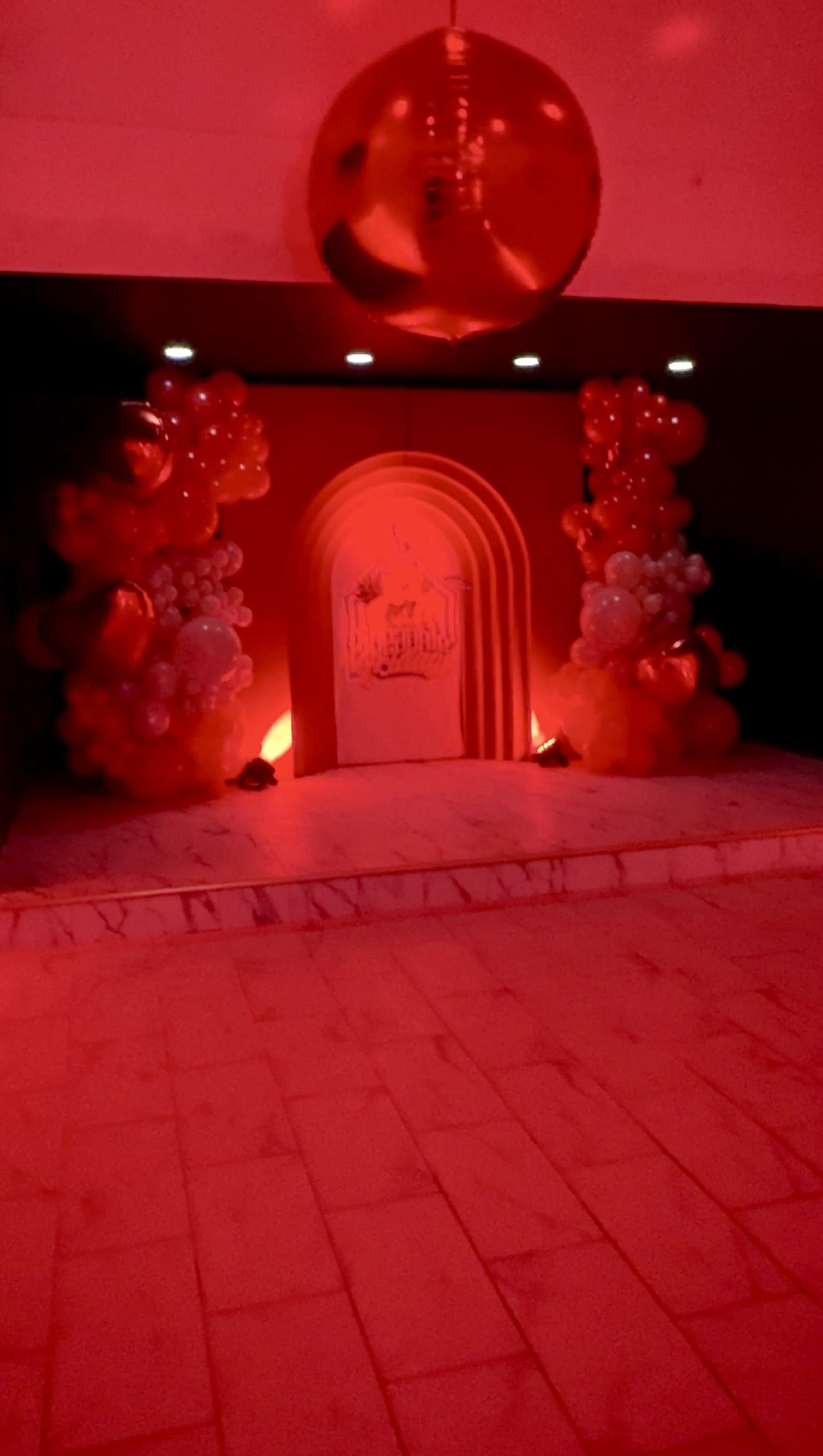 The image shows a decorated event space featuring an arch adorned with balloons and illuminated by red lighting.