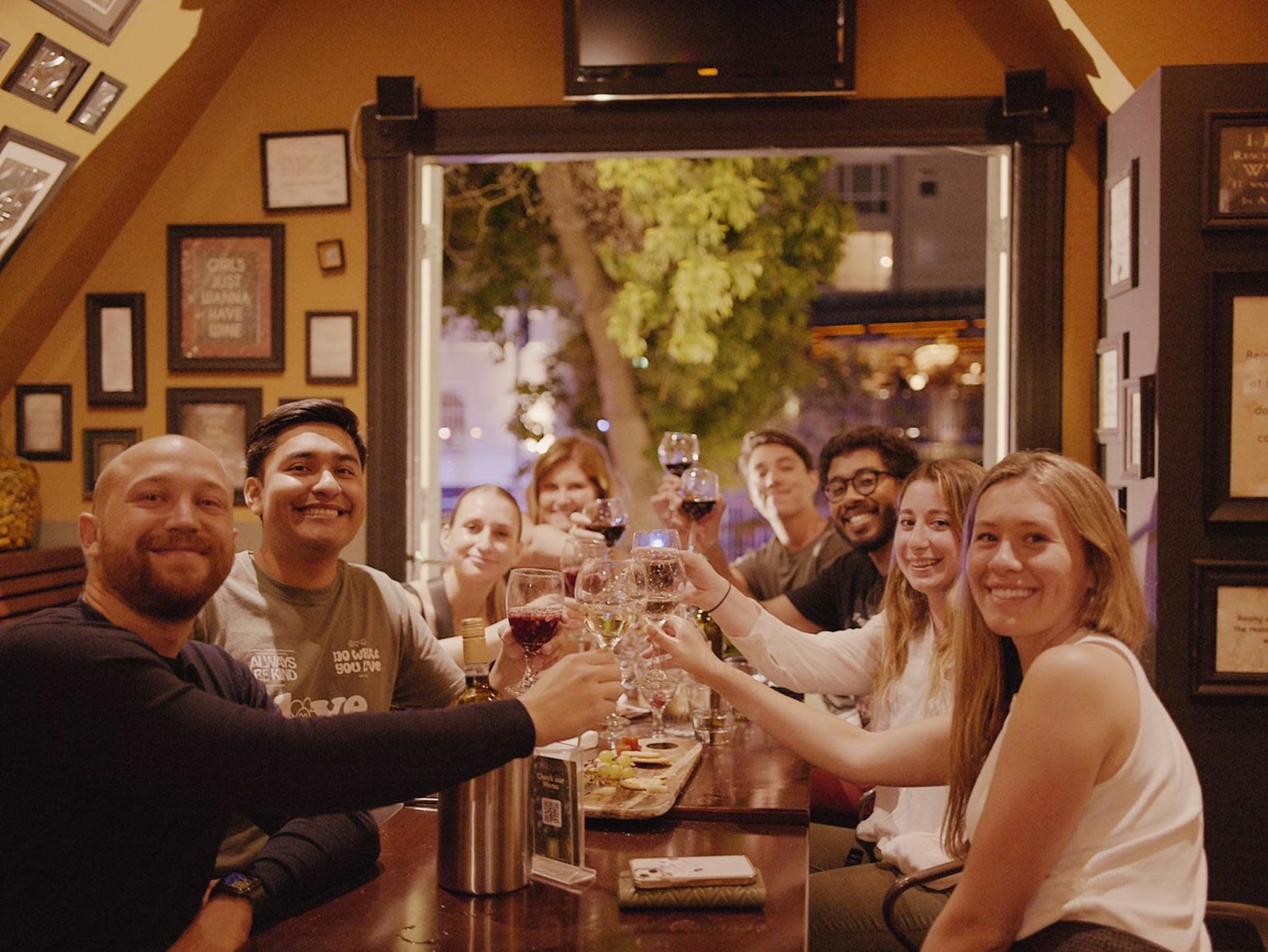 A group of people sit around a table in a warmly lit restaurant, smiling and raising their glasses in a toast.