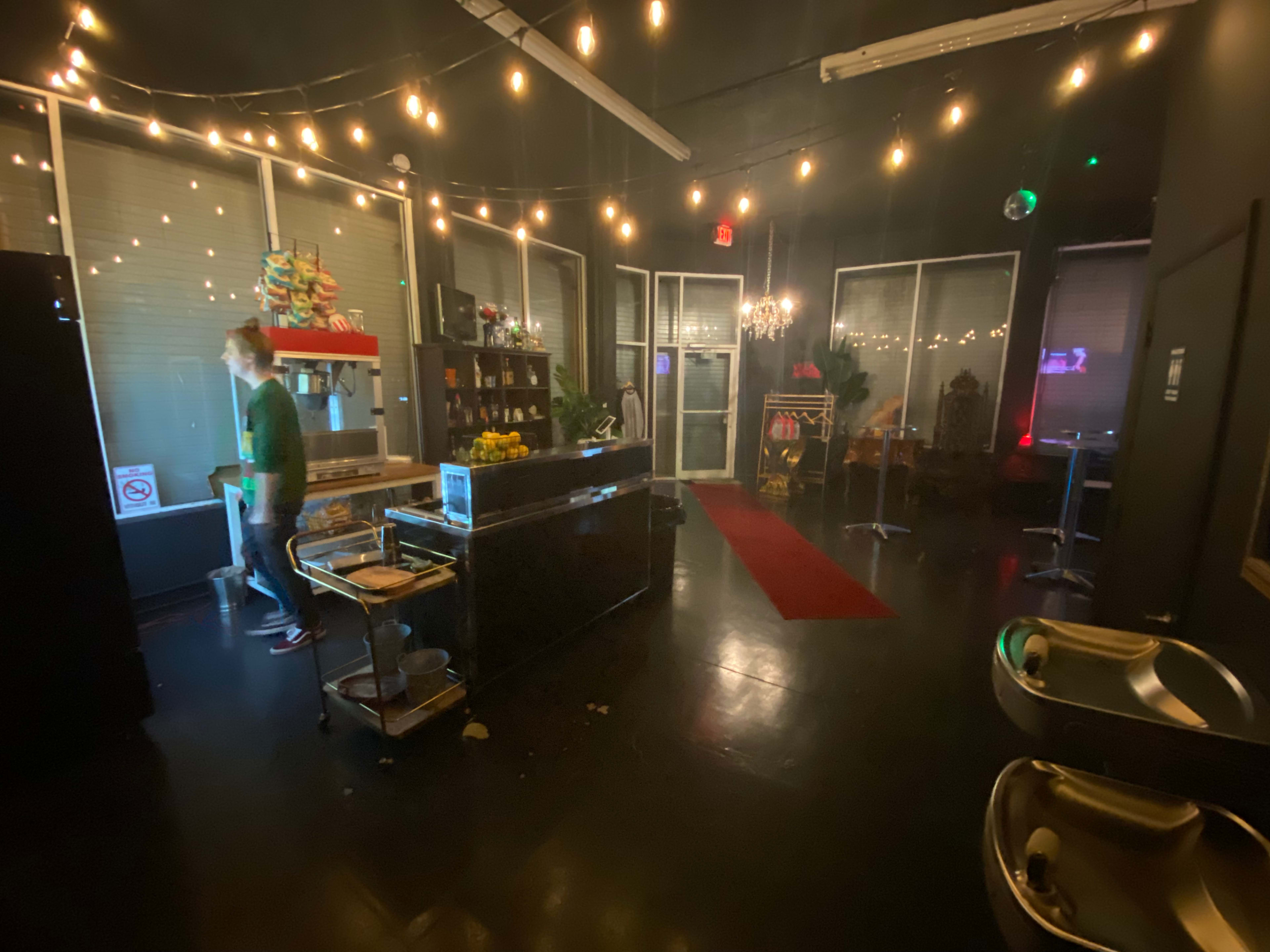 A dimly lit room with string lights, featuring a kitchen area on one side, a bar with high chairs, and a red carpet leading to a stage.