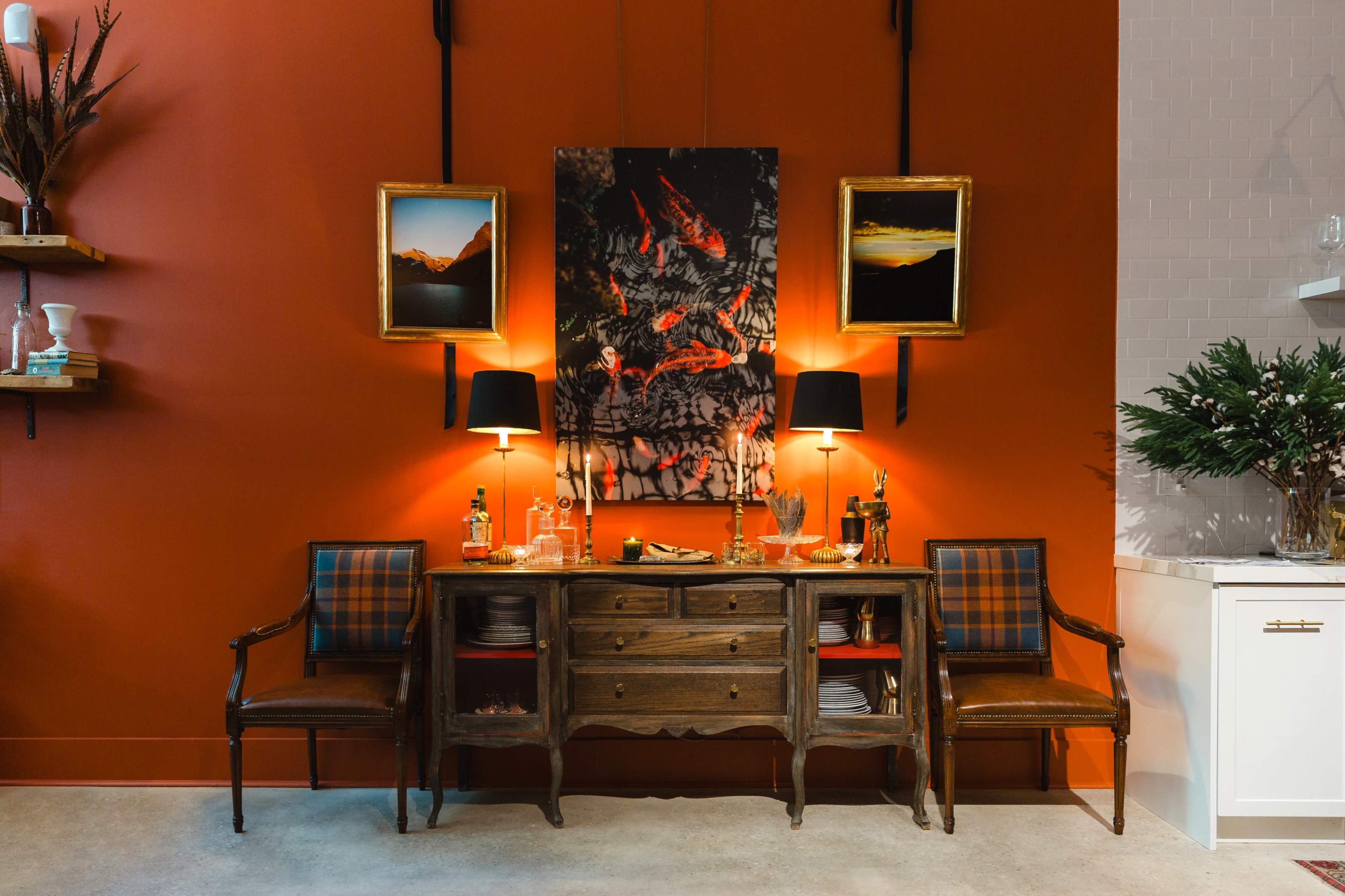 The image shows a wooden sideboard with two chairs and framed photographs hanging on an orange wall, accompanied by two table lamps and decorative items.