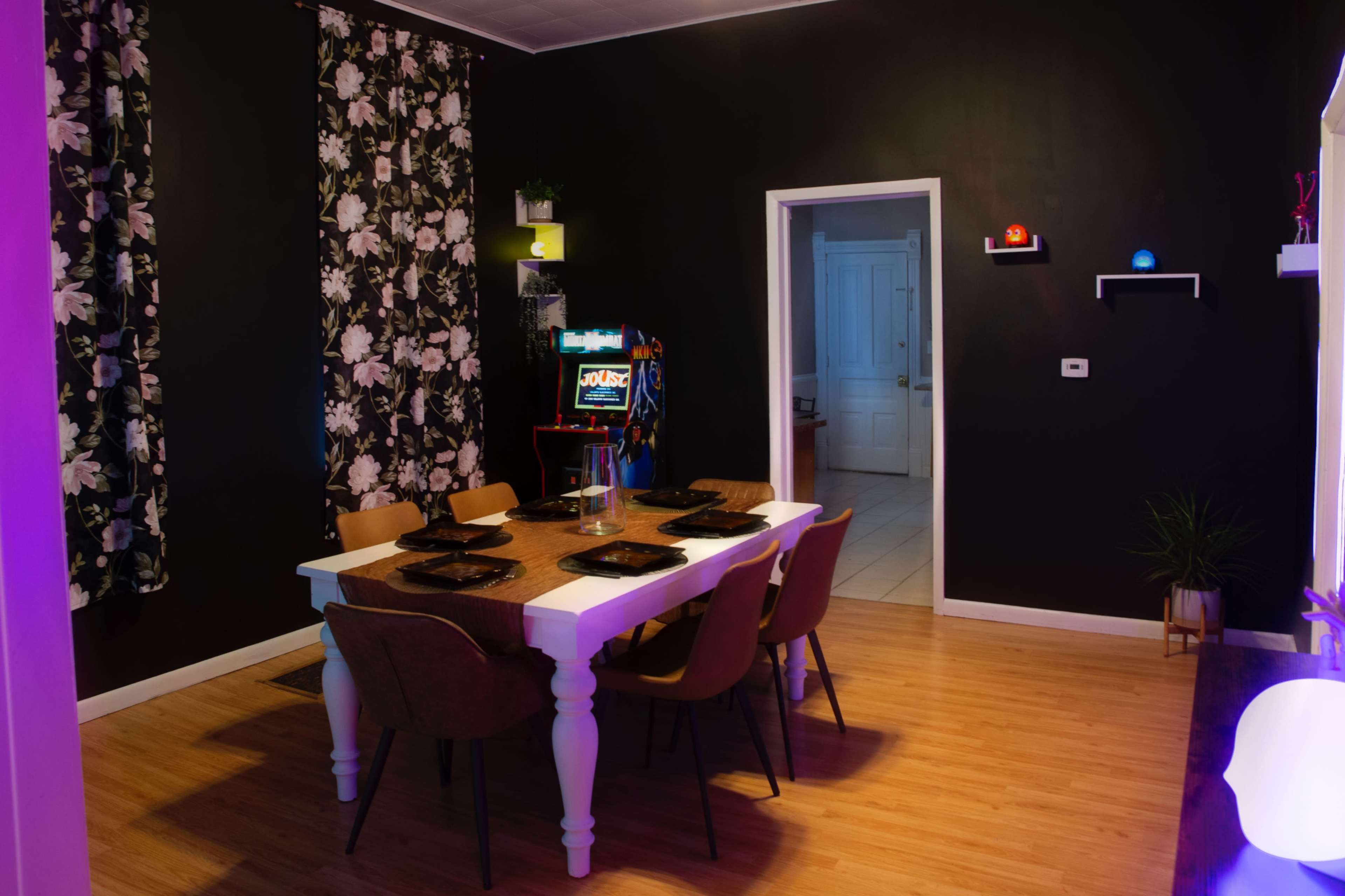 The image shows a dining room with a white table set for six, surrounded by wooden chairs, against a wall decorated with floral wallpaper and an arcade game machine in the background.
