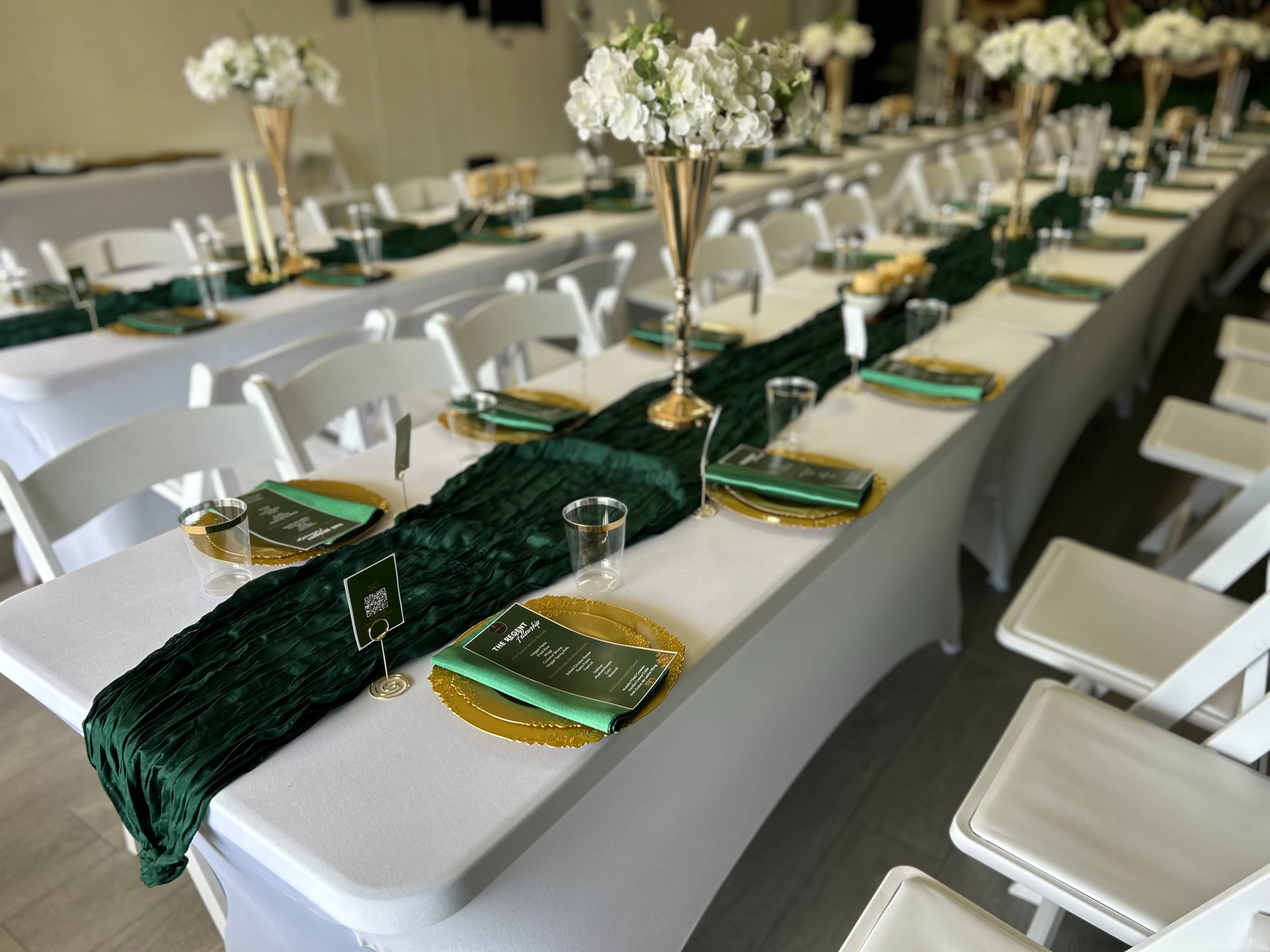 A banquet hall is set up with long tables covered in white linens, adorned with green table runners, gold charger plates, and floral centerpieces.