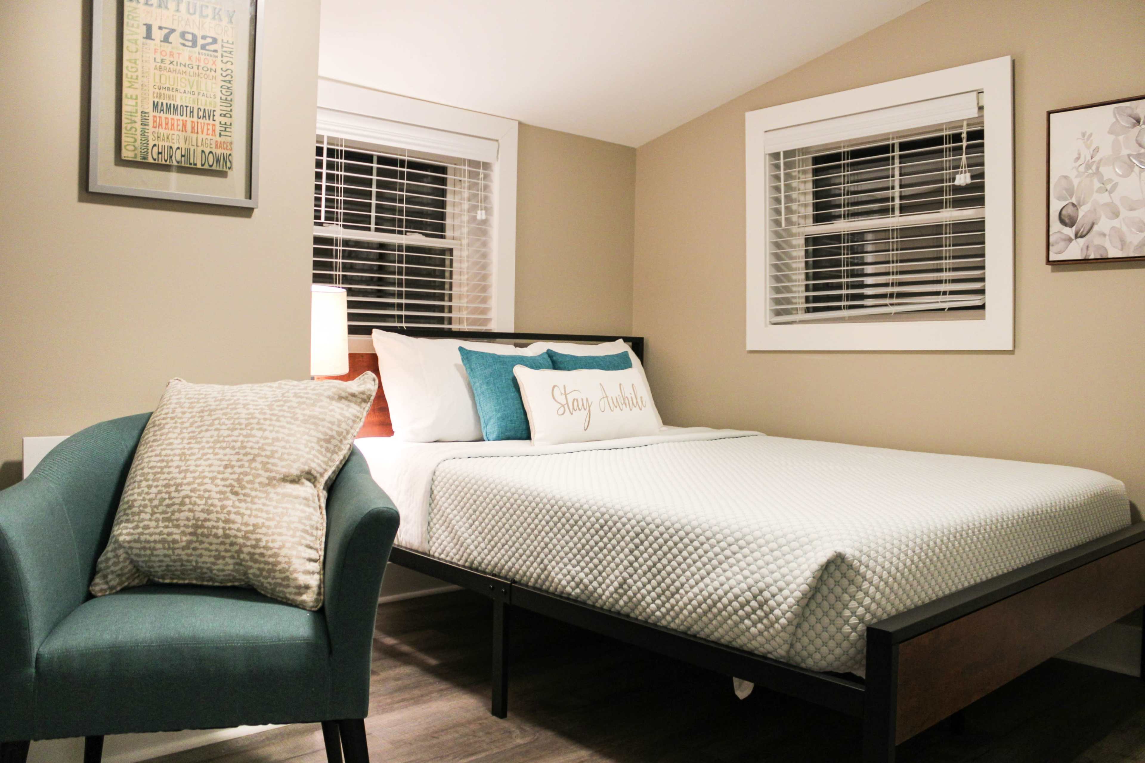 A bedroom with a bed, a light blue accent chair, and two windows, featuring beige walls and wood flooring.