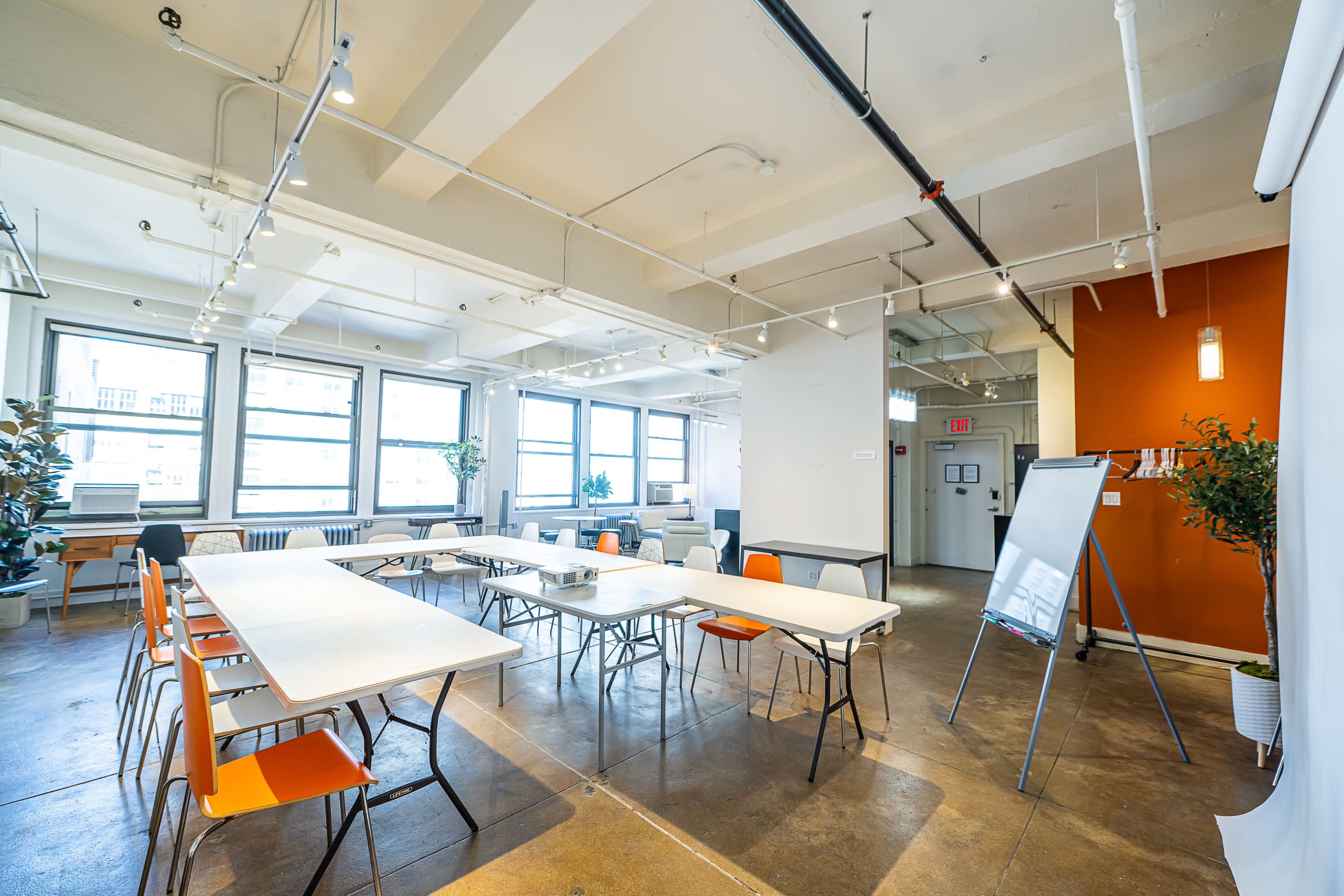 The image shows a bright, modern meeting room with several tables arranged in a U-shape, surrounded by chairs, large windows, and an accent wall in orange.