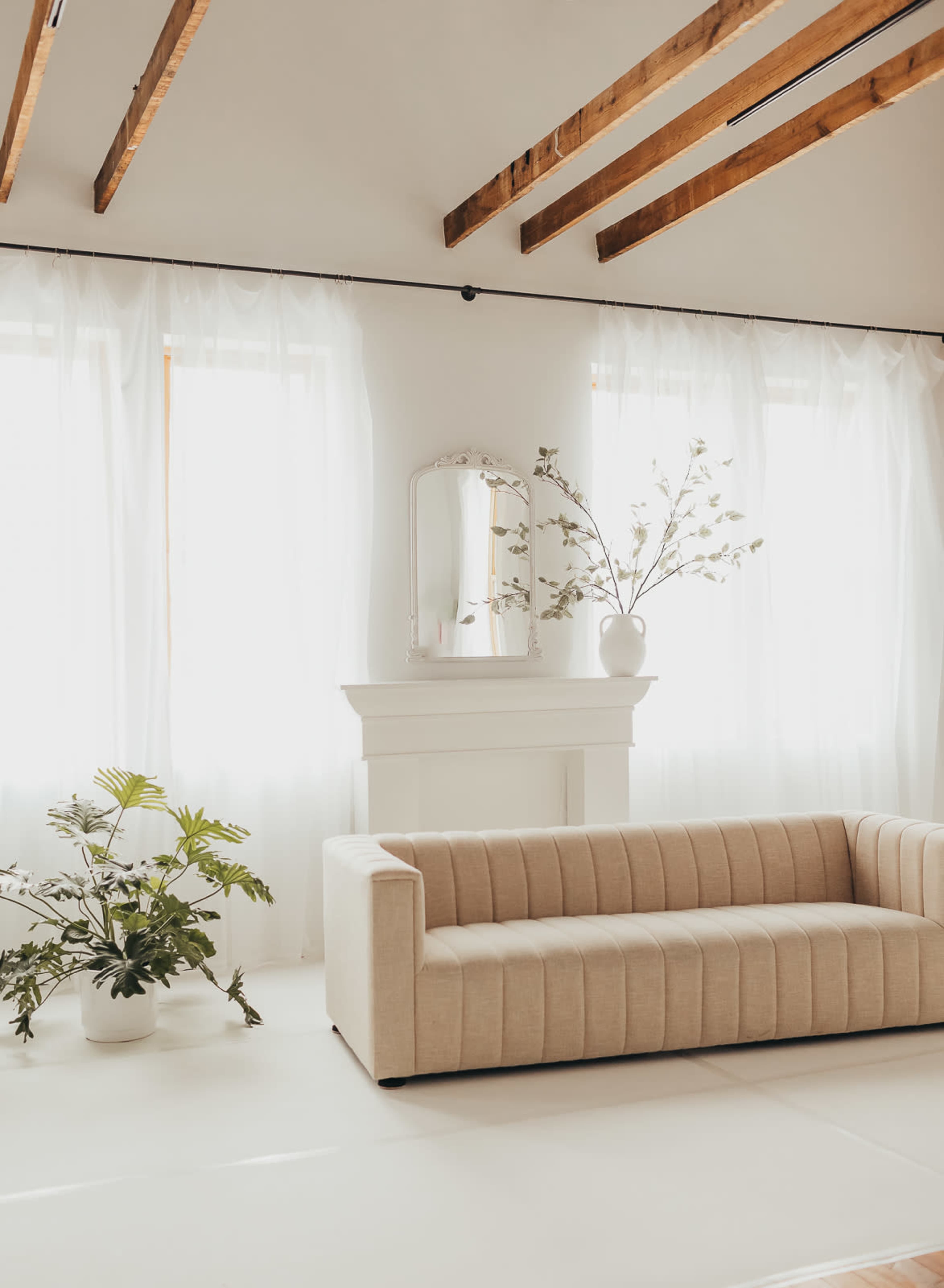 A beige, tufted sofa sits in a bright room with large windows draped in sheer curtains, beside potted plants and a decorative mirror on a white mantel.