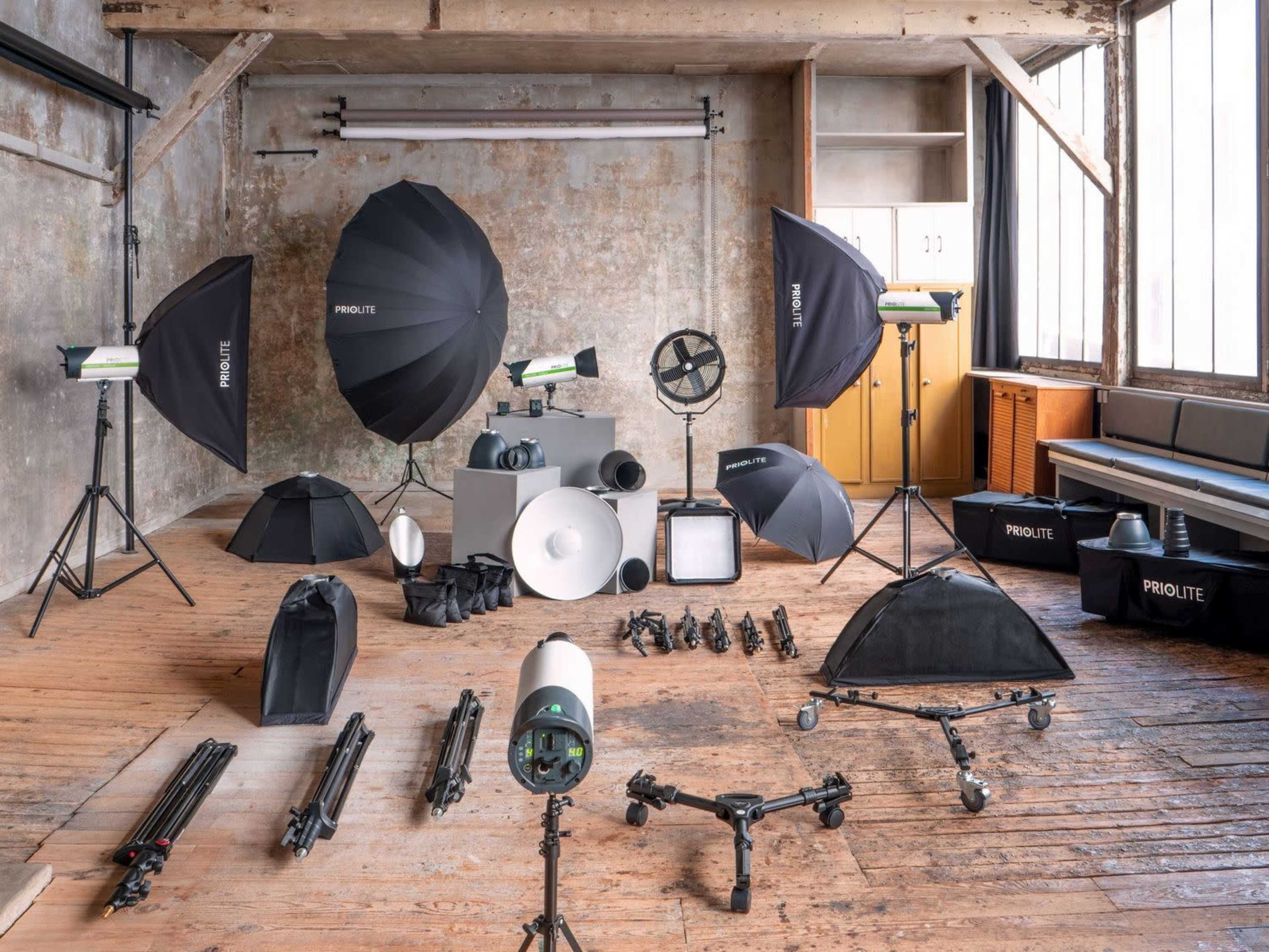 The image shows a photography studio setup featuring various lighting equipment, softboxes, tripods, and accessories arranged in a spacious room with wooden floors and large windows.