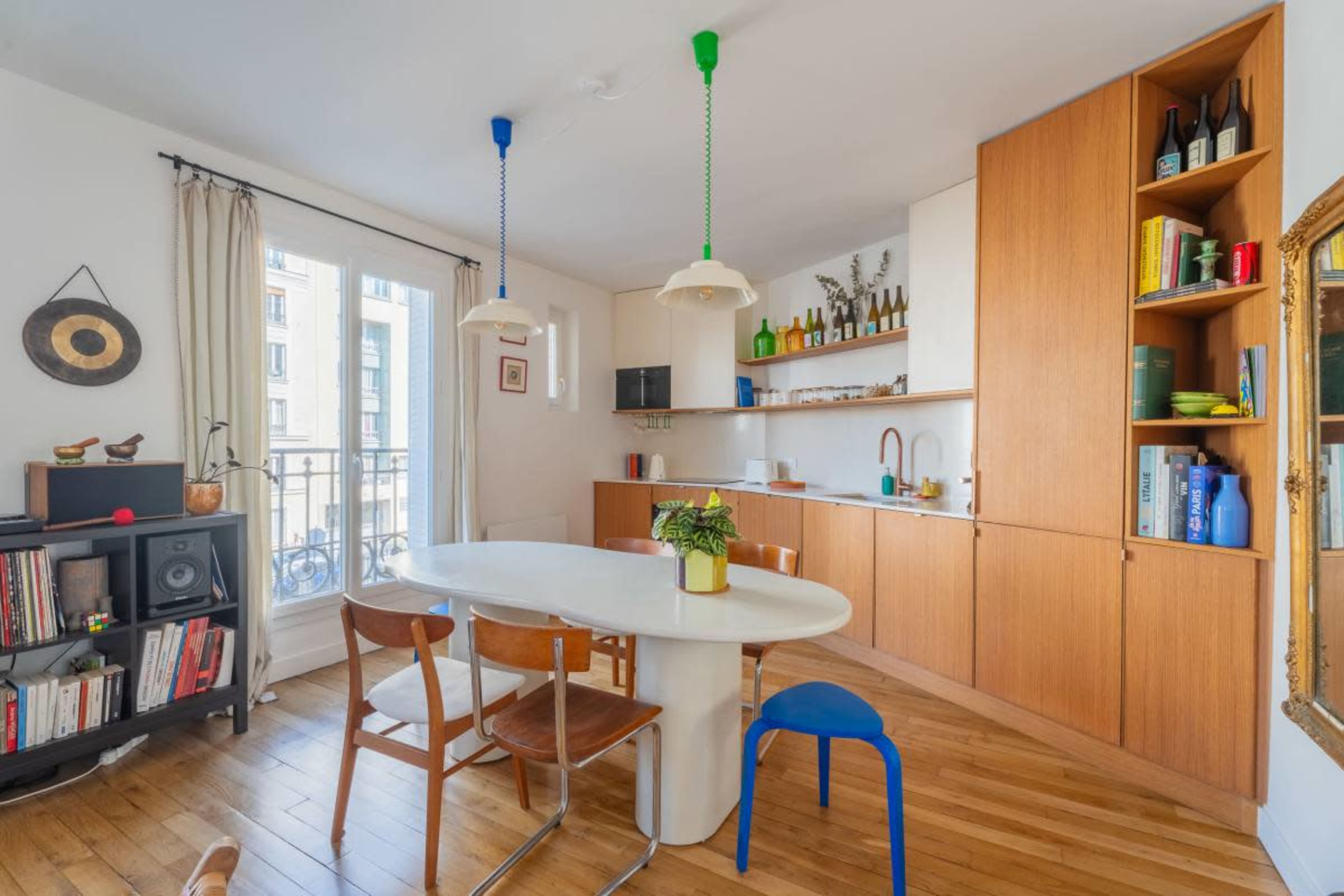 The image shows a modern kitchen and dining area featuring a round table with wooden chairs, cabinetry, and colorful hanging lights.
