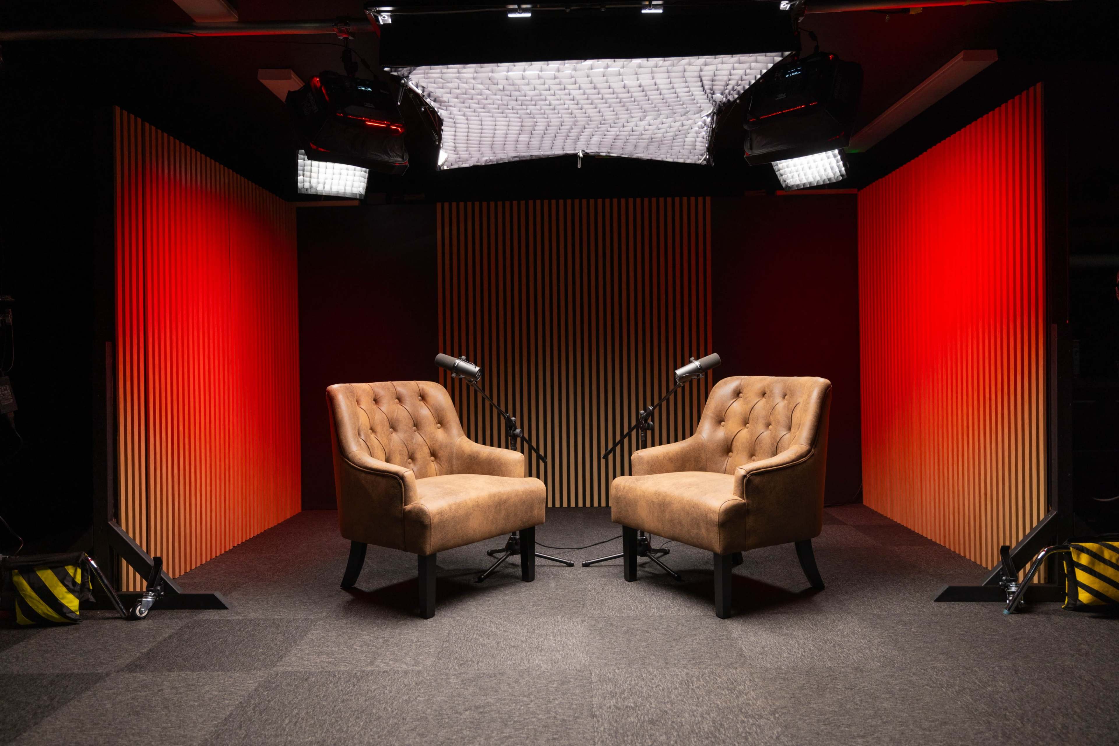 The image shows two brown tufted chairs facing each other in a studio setting, with two microphones positioned nearby and bright lighting highlighting the wooden panel backdrop.
