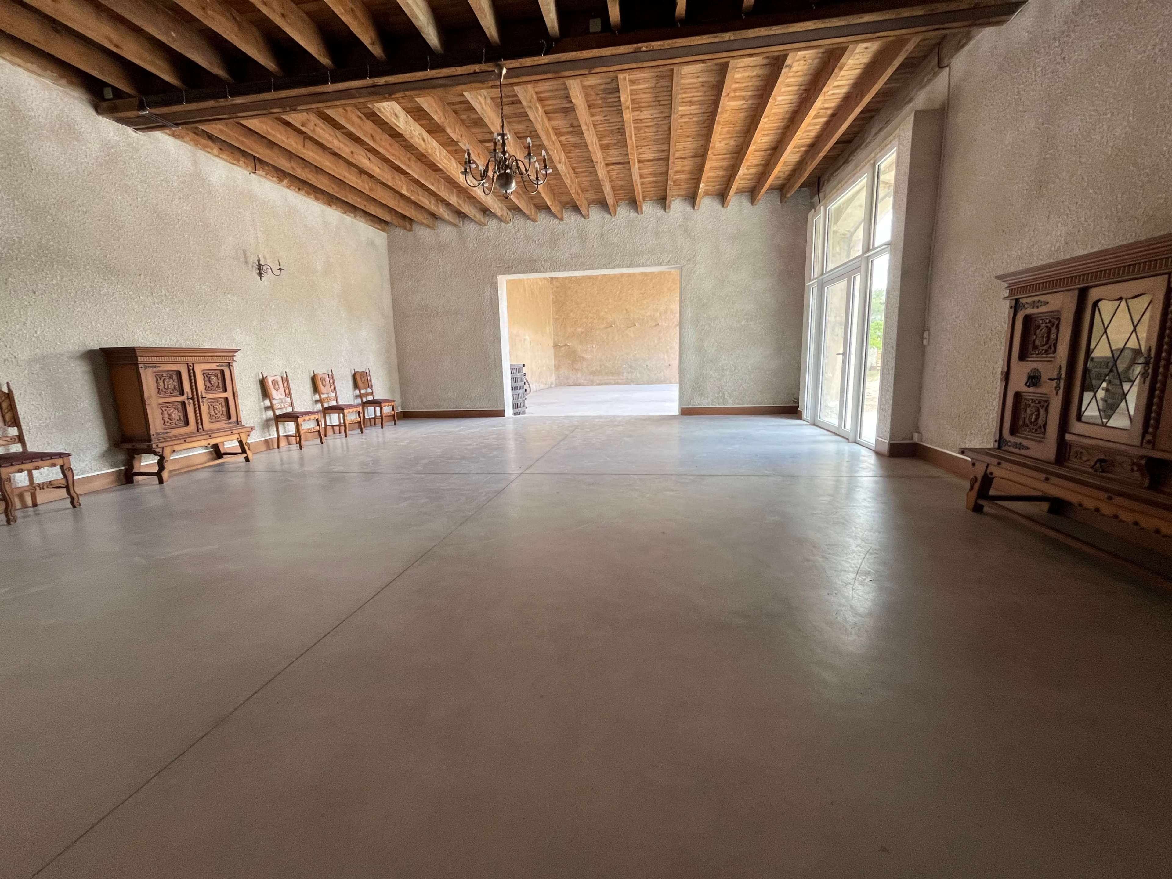 The image shows a spacious room with high wooden beams, a polished concrete floor, and antique wooden furniture along the walls.