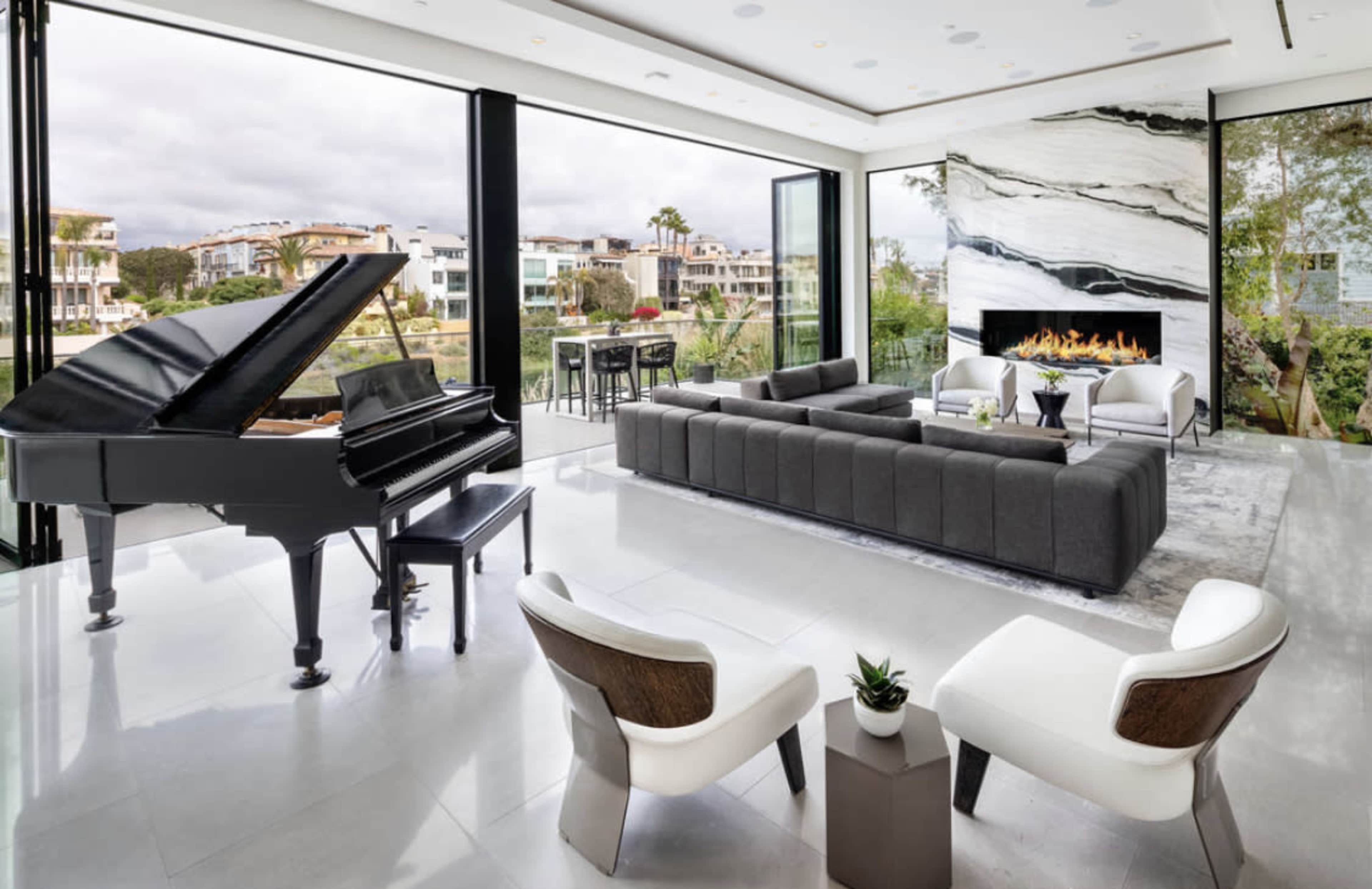 A modern living room featuring a grand piano, a sectional sofa, two white chairs, and large glass doors opening to an outdoor view with a fireplace and greenery.