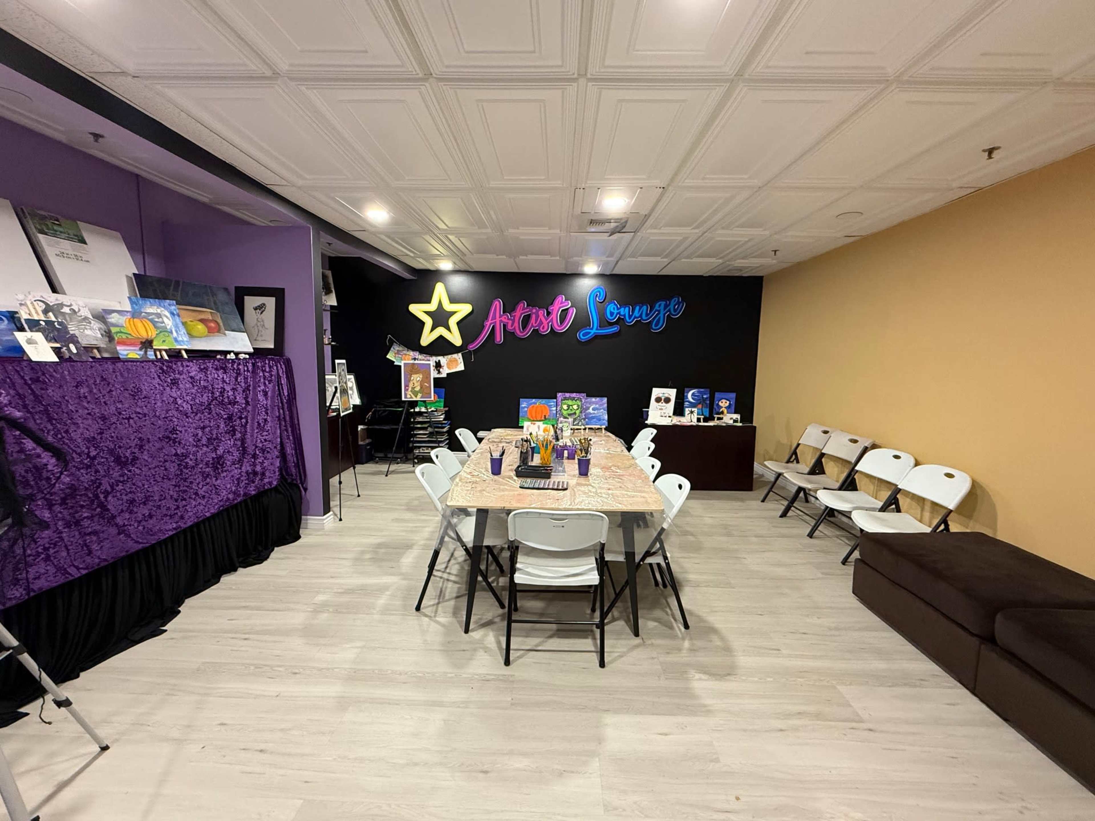 The image shows an art studio with a decorated table in the center, surrounded by chairs, and walls featuring vibrant colors and artwork displays.