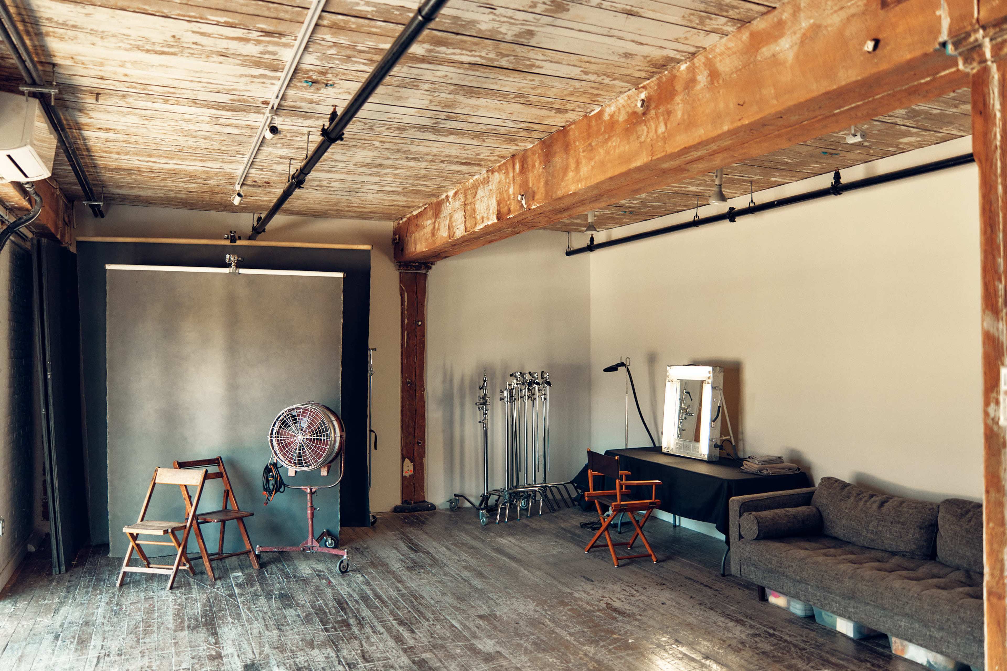 The image shows a spacious photo studio with a wooden floor, two chairs, a large fan, a makeup station, and backdrop on one side.