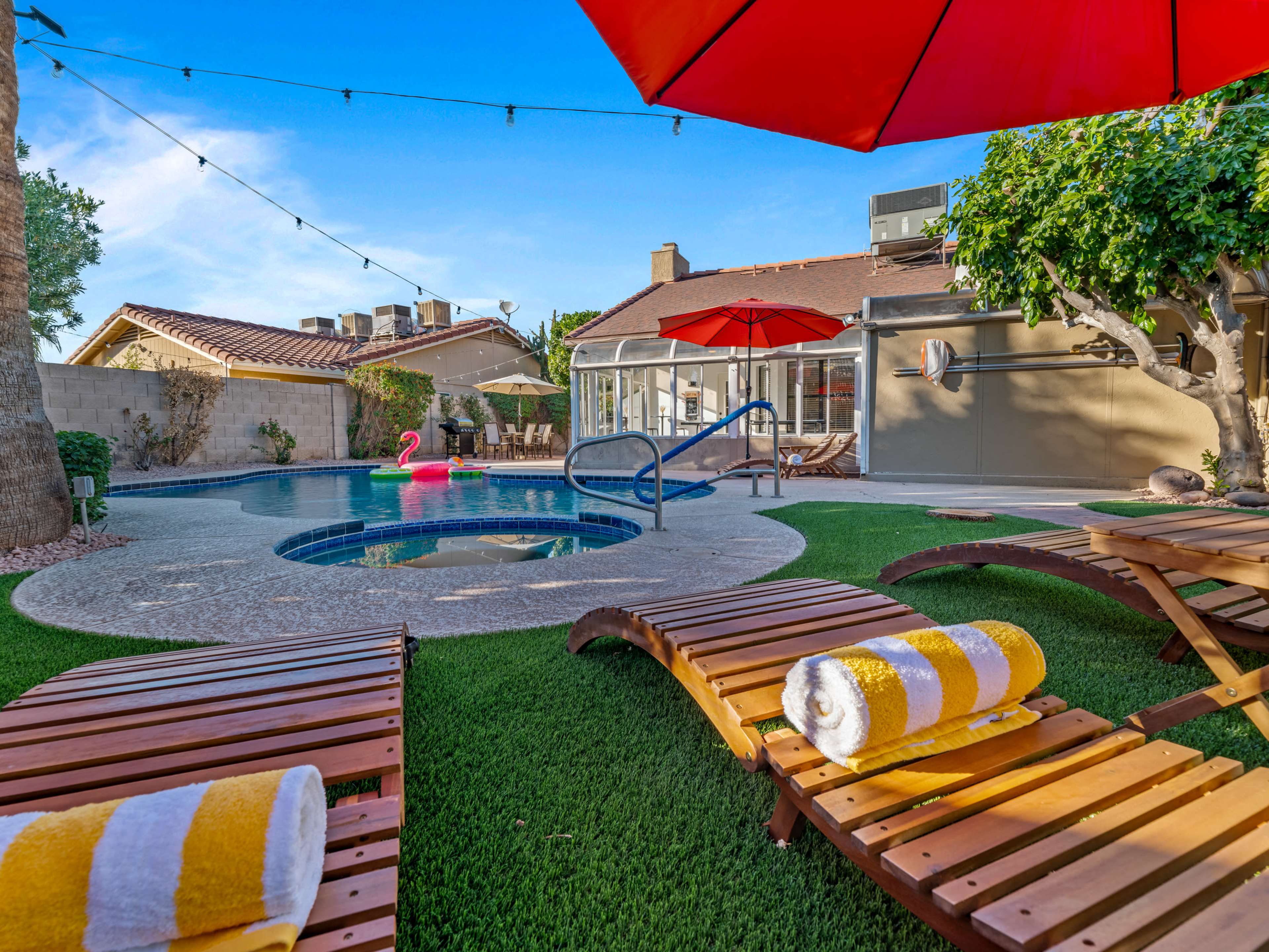 A backyard features a pool surrounded by lounge chairs, an umbrella, and a pink inflatable flamingo.