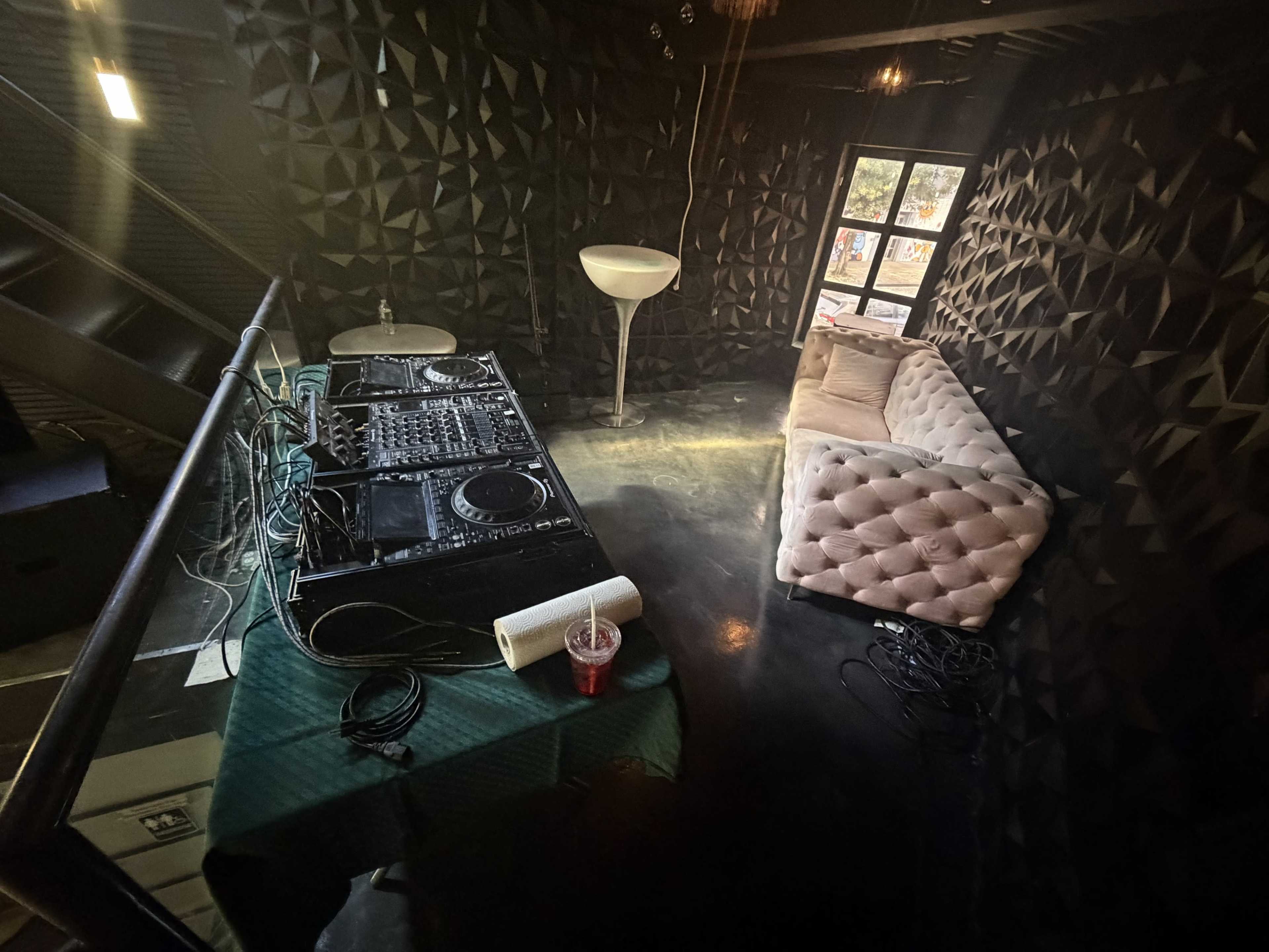 The image shows a dimly lit room with a DJ setup featuring turntables and a mixer, accompanied by a plush sofa and a lamp against a textured black wall.