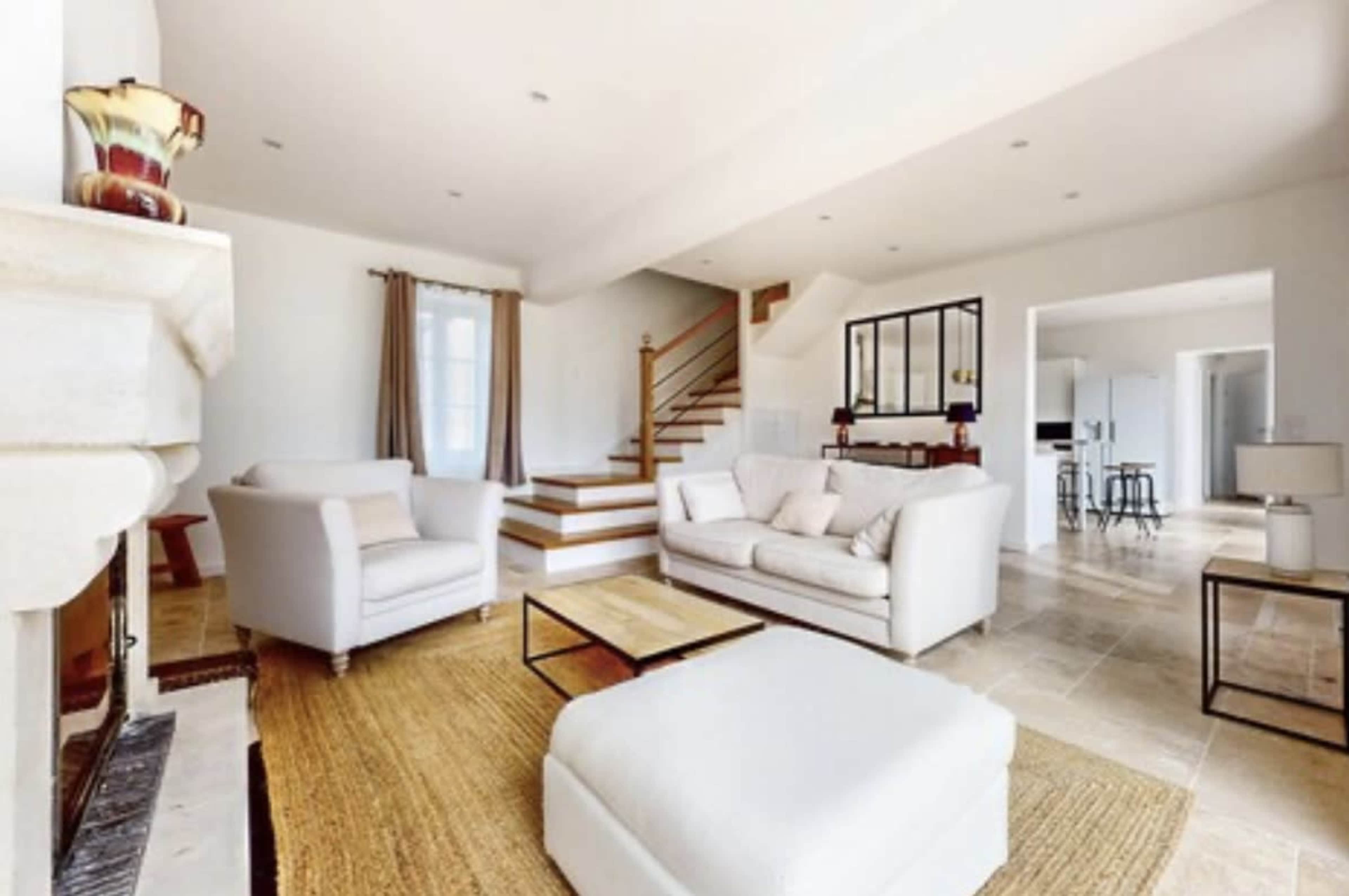 The image shows a spacious living room with white sofas, a coffee table, a staircase, and a light-colored rug on a stone floor.