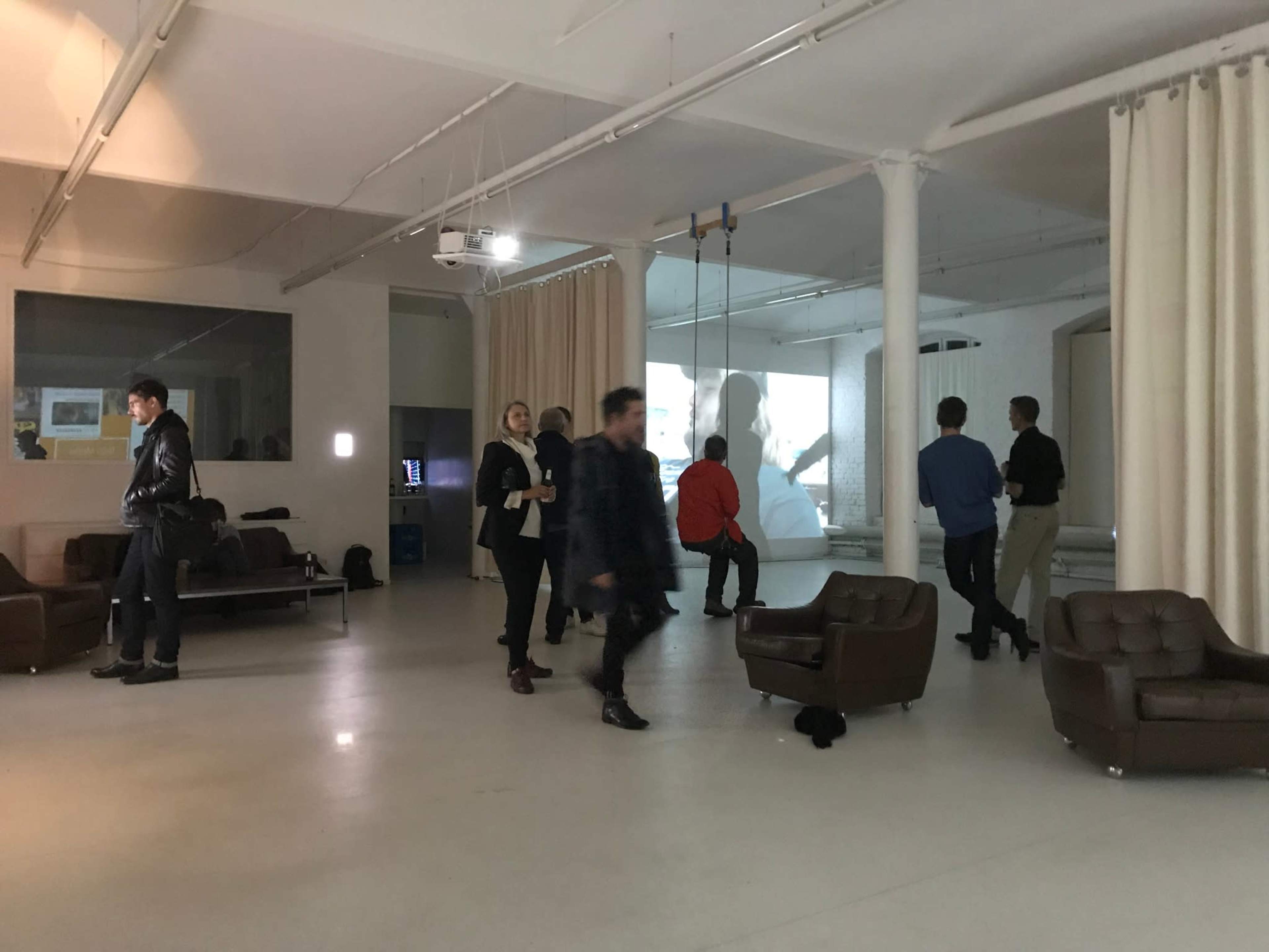 A spacious, minimalistic gallery with people interacting and viewing a projected video installation, while some are seated on leather couches.