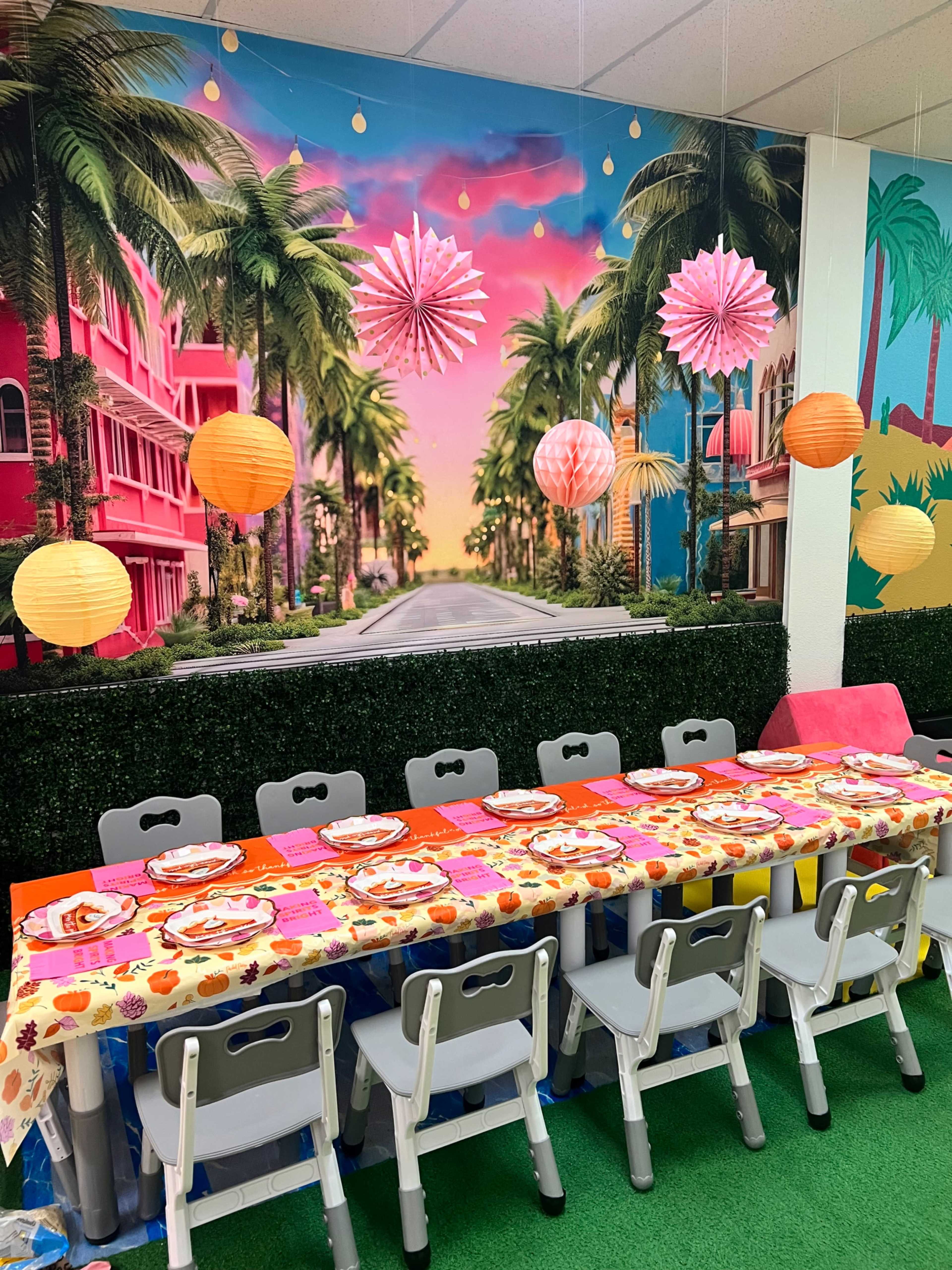 A long table set with colorful tableware and surrounded by chairs is decorated with paper lanterns and a vibrant mural of a palm-lined street.