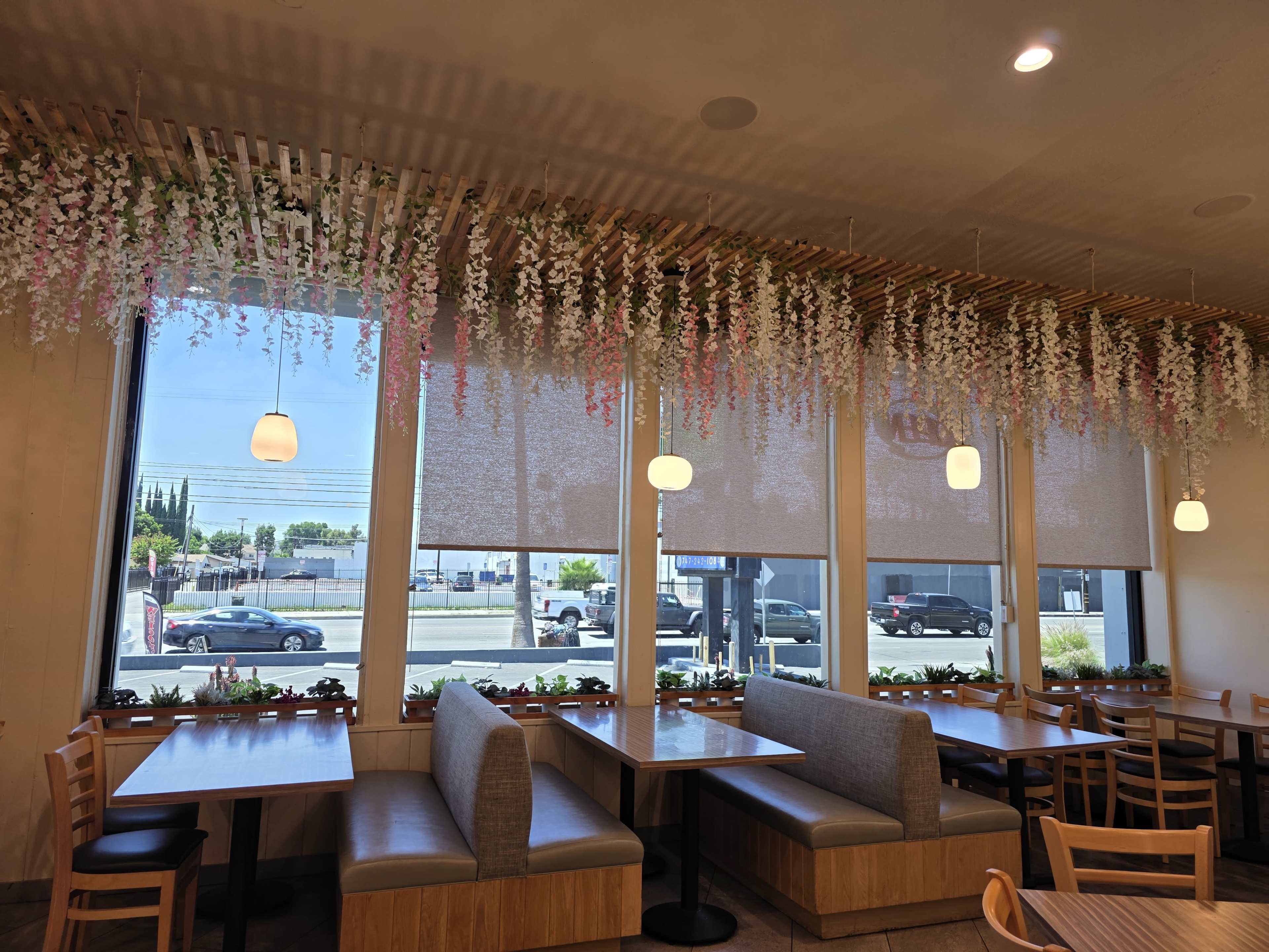 The image shows a restaurant interior with wooden tables and benches, large windows adorned with floral decorations, and light fixtures hanging from the ceiling.
