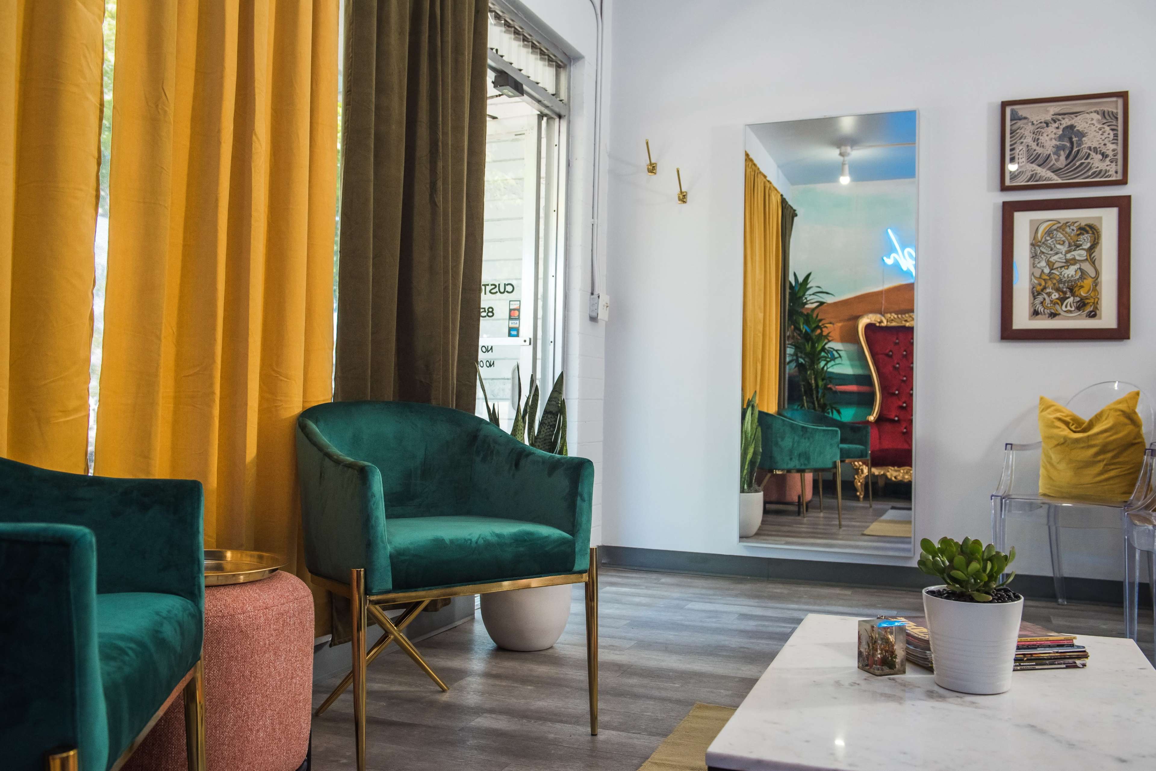 The image shows a brightly lit room featuring green velvet chairs, yellow and green curtains, a large mirror, and a small coffee table with decorative items.