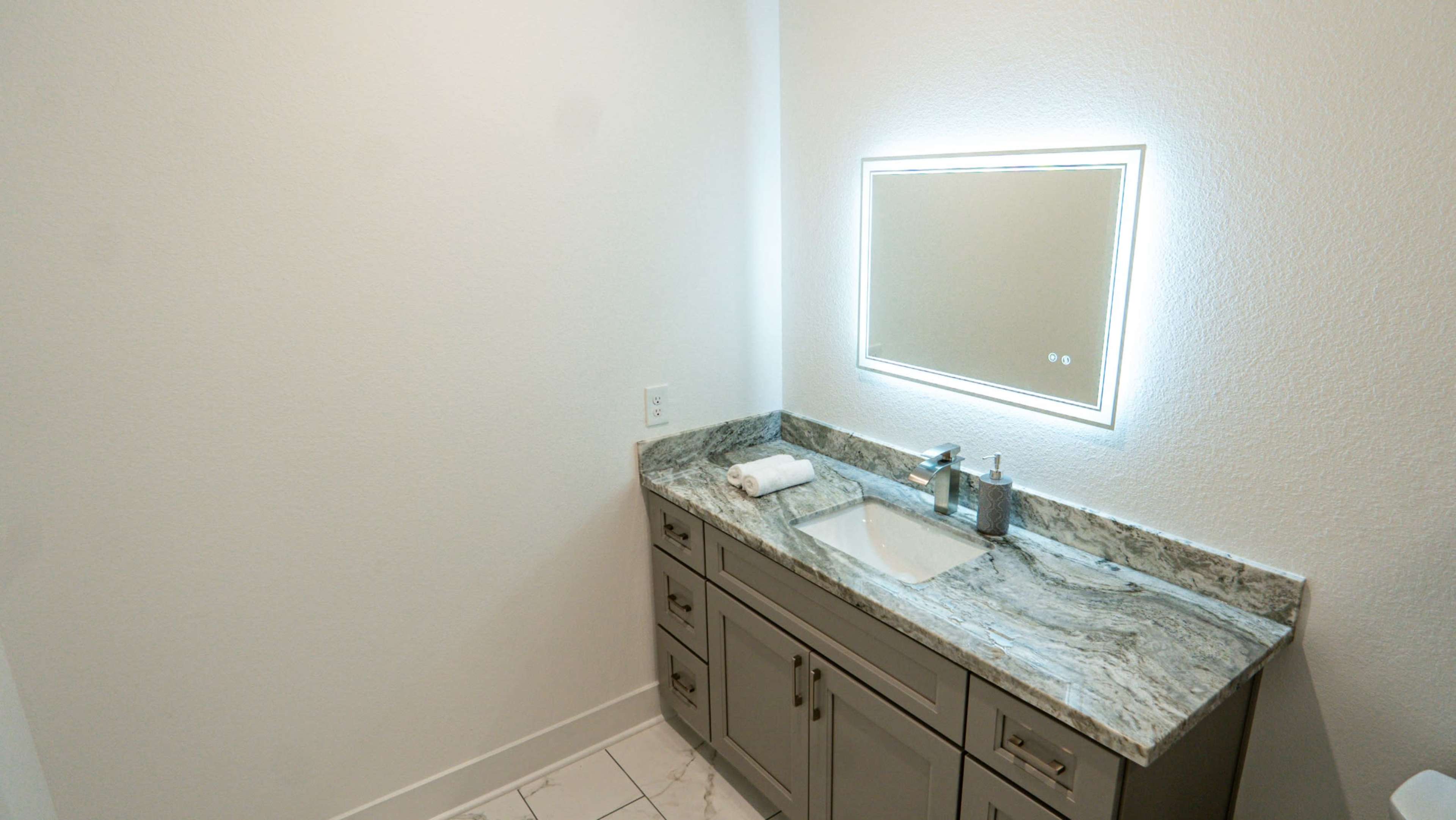 The image shows a modern bathroom with a granite countertop, a mirror with built-in lighting, and a sink.