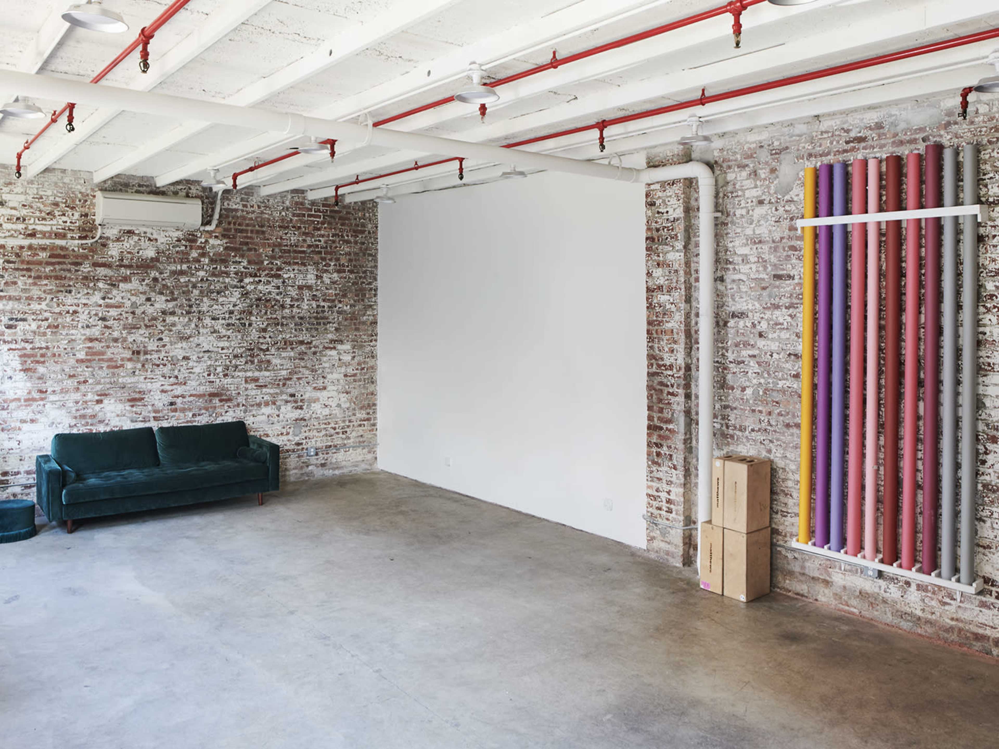 The image features an empty industrial-style room with exposed brick walls, polished concrete floors, a green sofa, and colorful vertical pipes on one side.