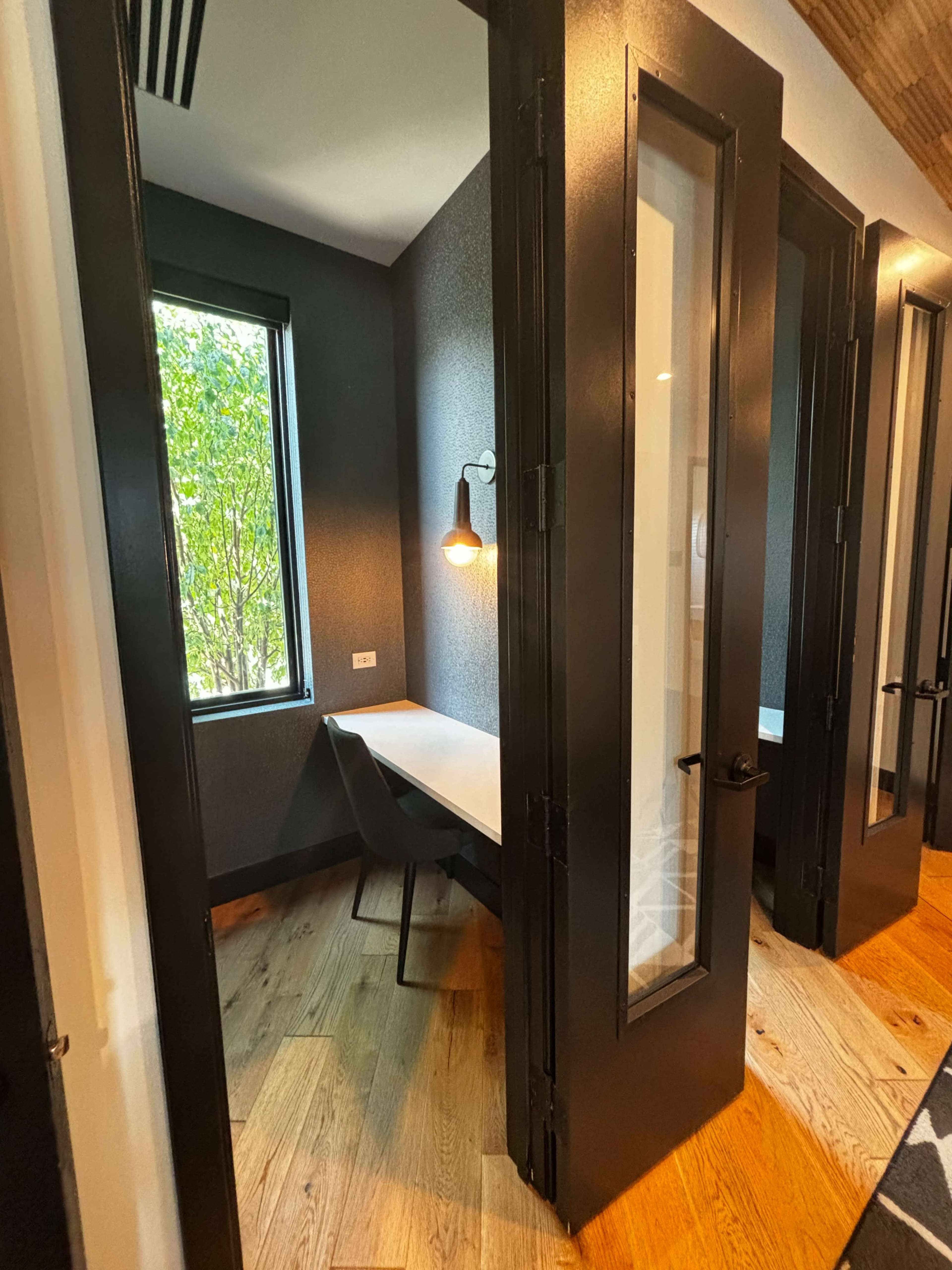 A small, enclosed workspace with a single desk and a chair, featuring large black doors and a window allowing natural light to enter.