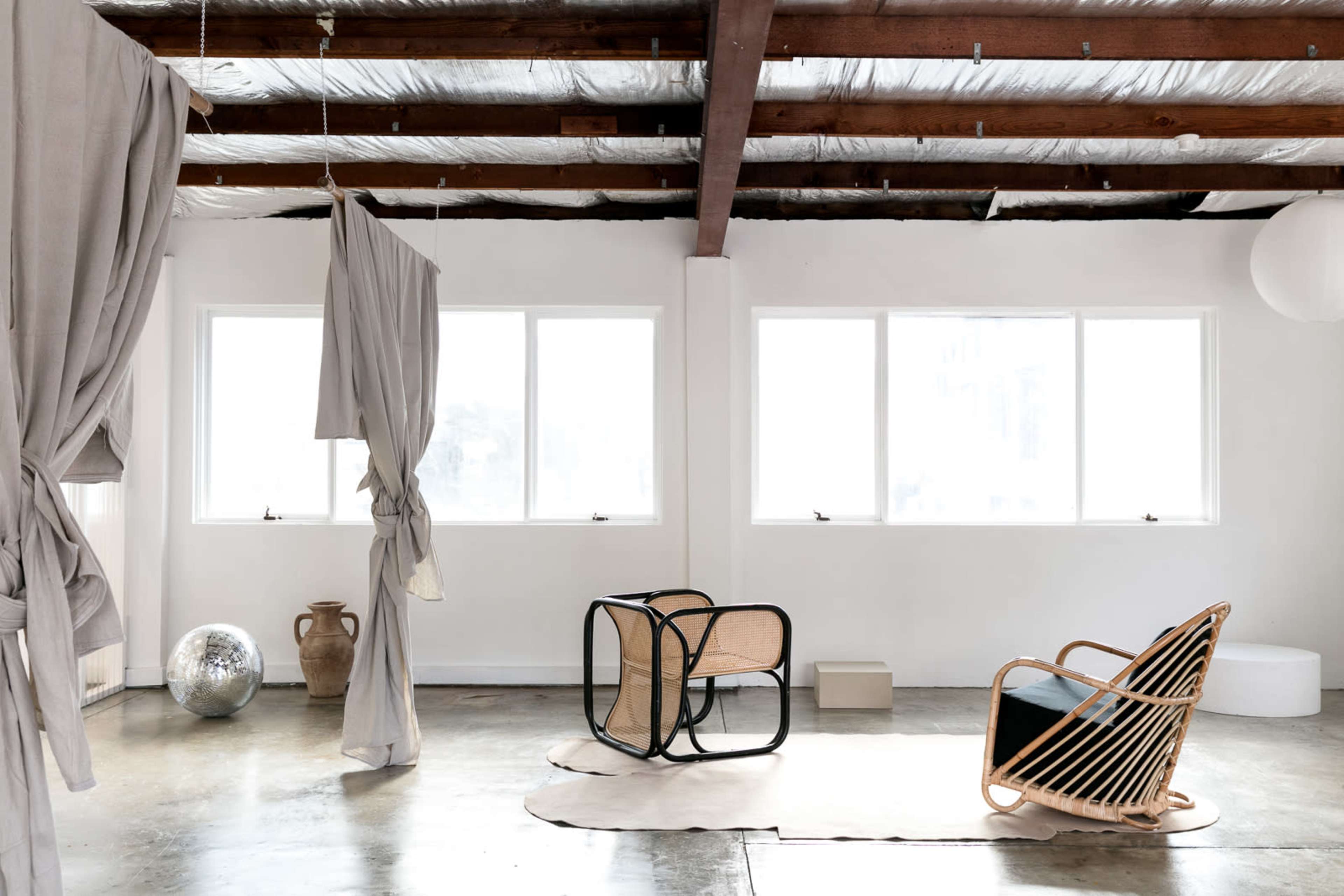 A spacious room features large windows, a woven chair, and minimal furnishings against a backdrop of exposed wooden beams and concrete flooring.