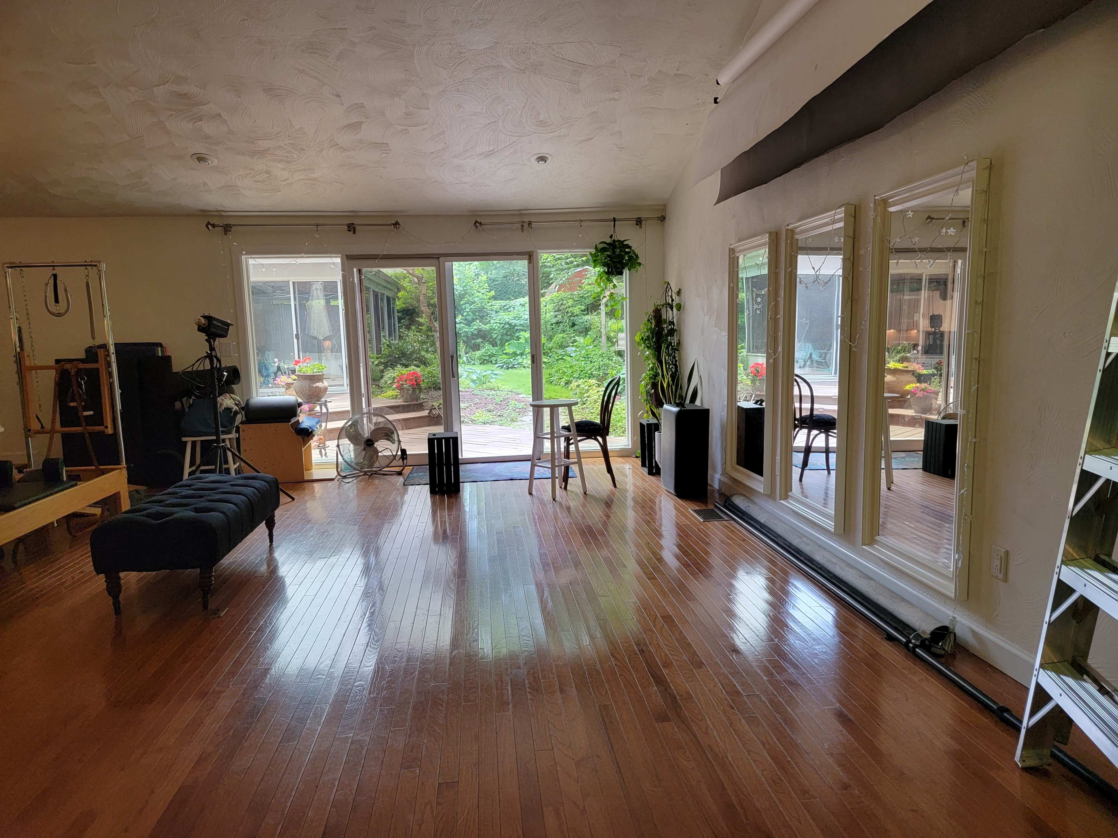 The image shows a spacious room with wooden floors, large mirrors on one wall, and a view of a garden through sliding glass doors.