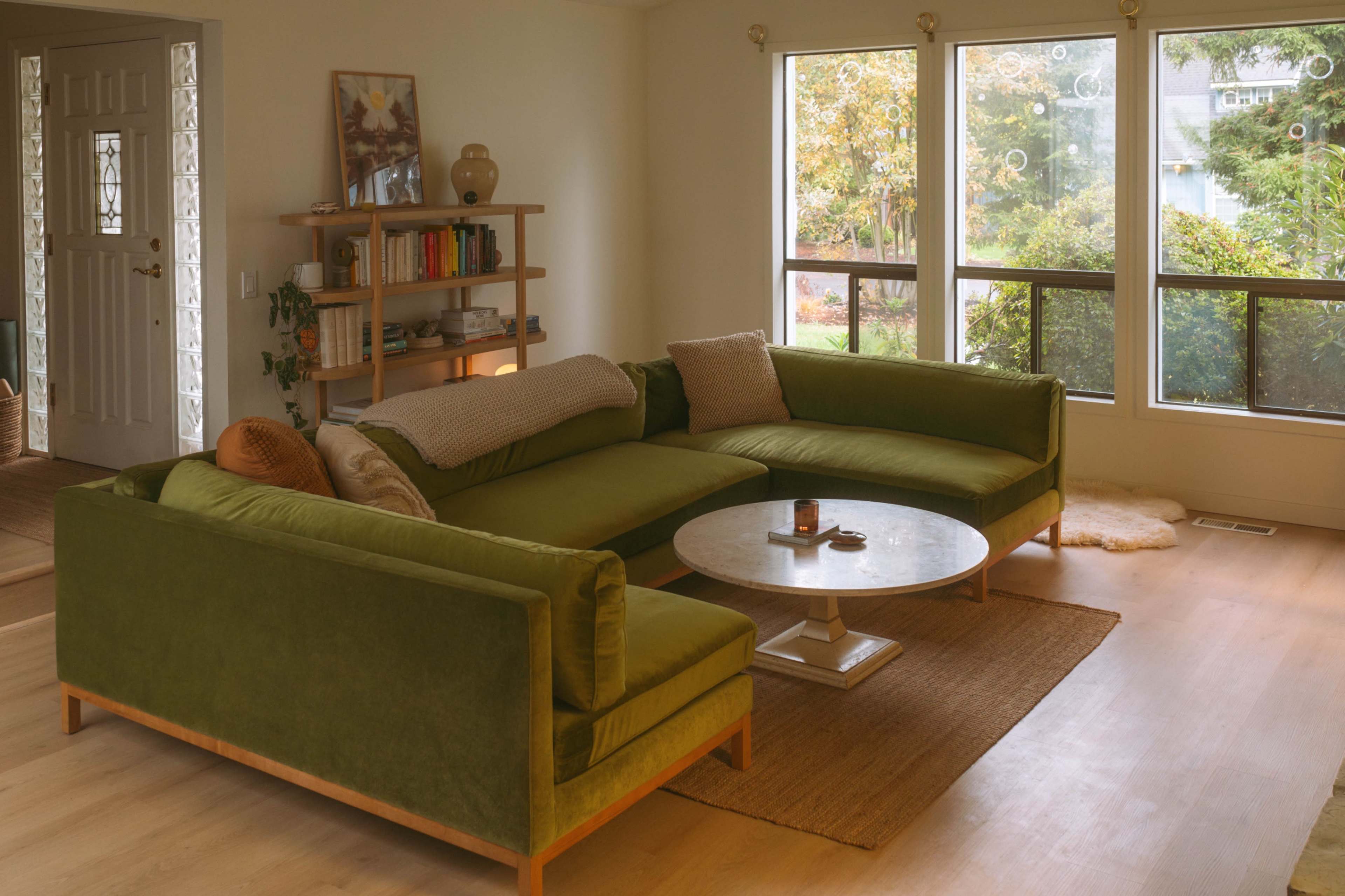 The image shows a cozy living room with a green sectional sofa, a round coffee table, large windows providing natural light, and plants visible outside.