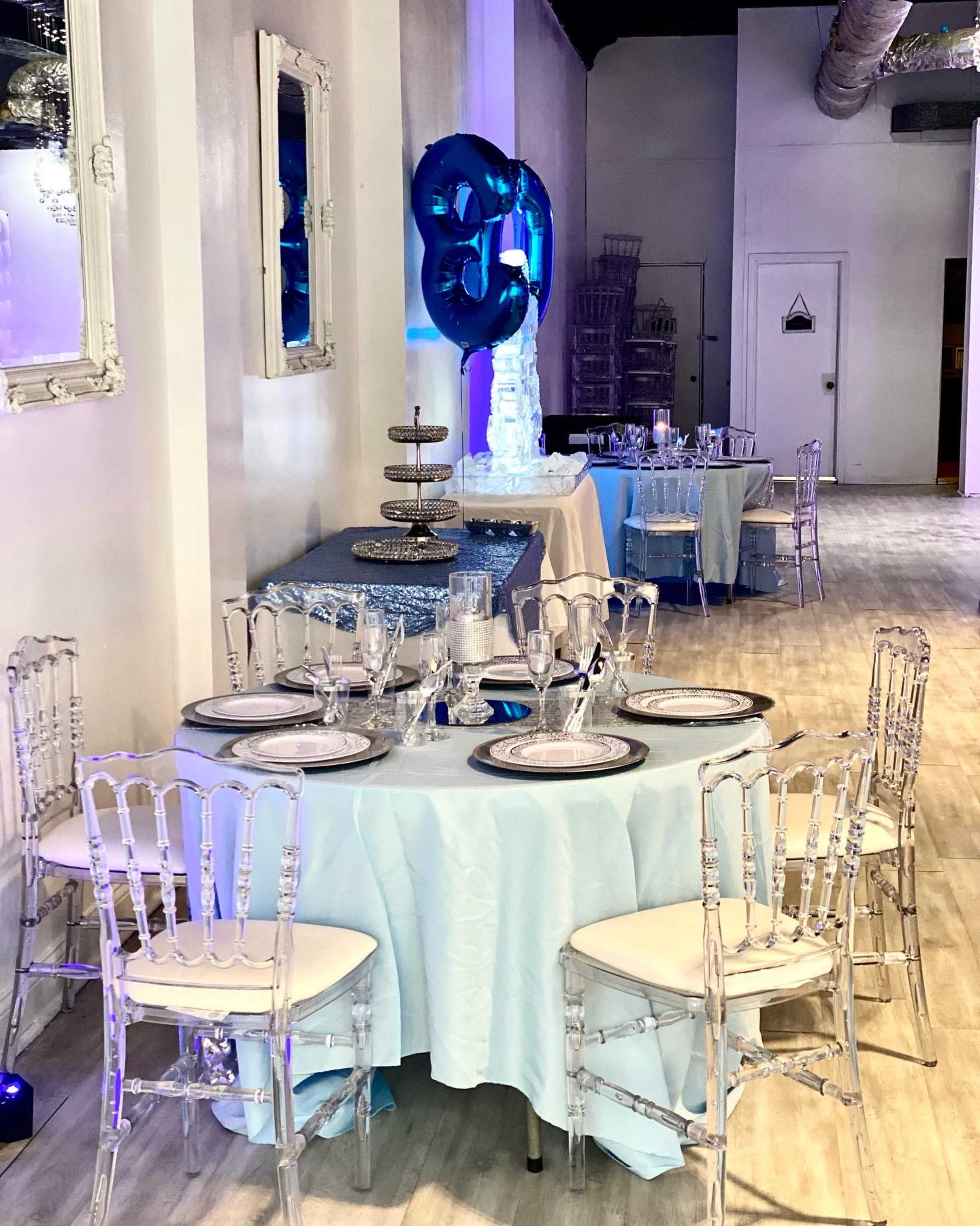 A banquet hall with elegantly set tables featuring light blue tablecloths, clear chiavari chairs, and a decorated backdrop with large number balloons.