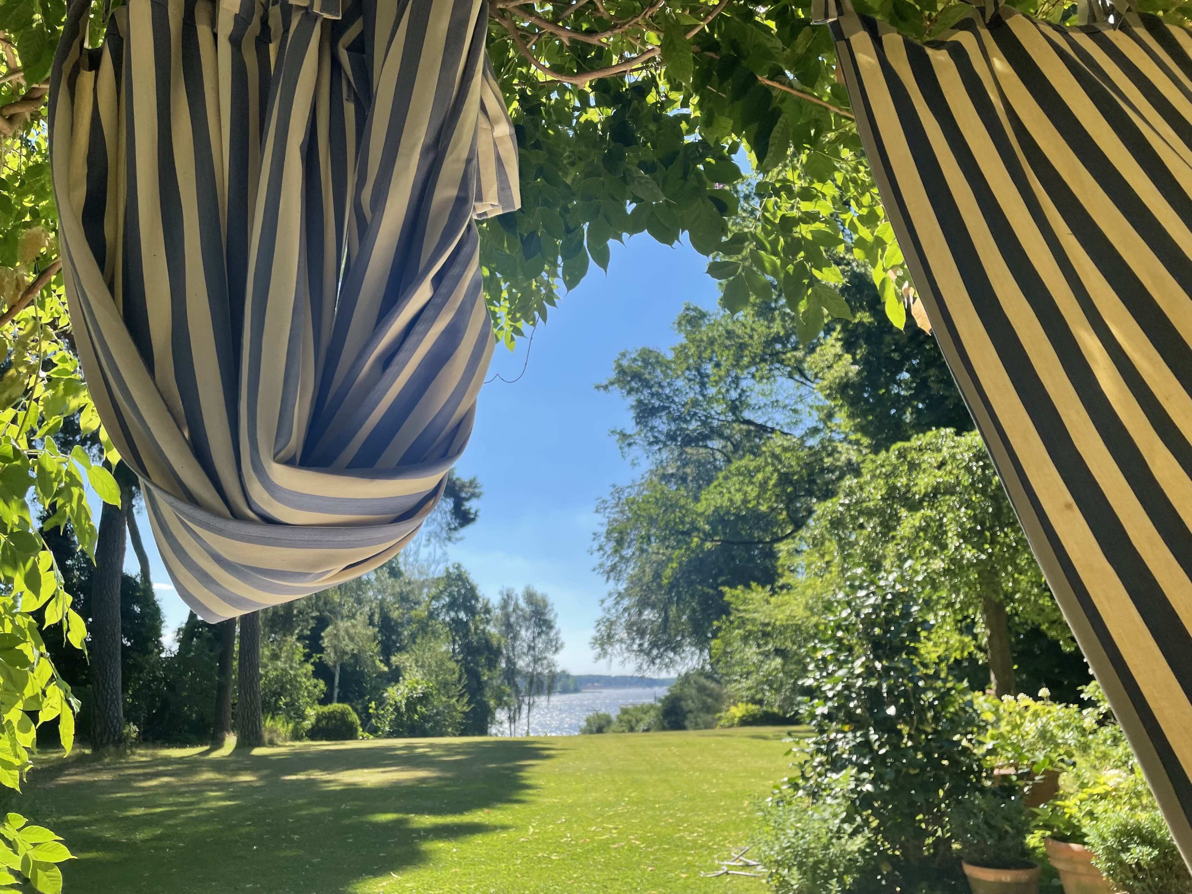 A view through striped fabric drapes reveals a sunny garden with trees and a lake in the background.