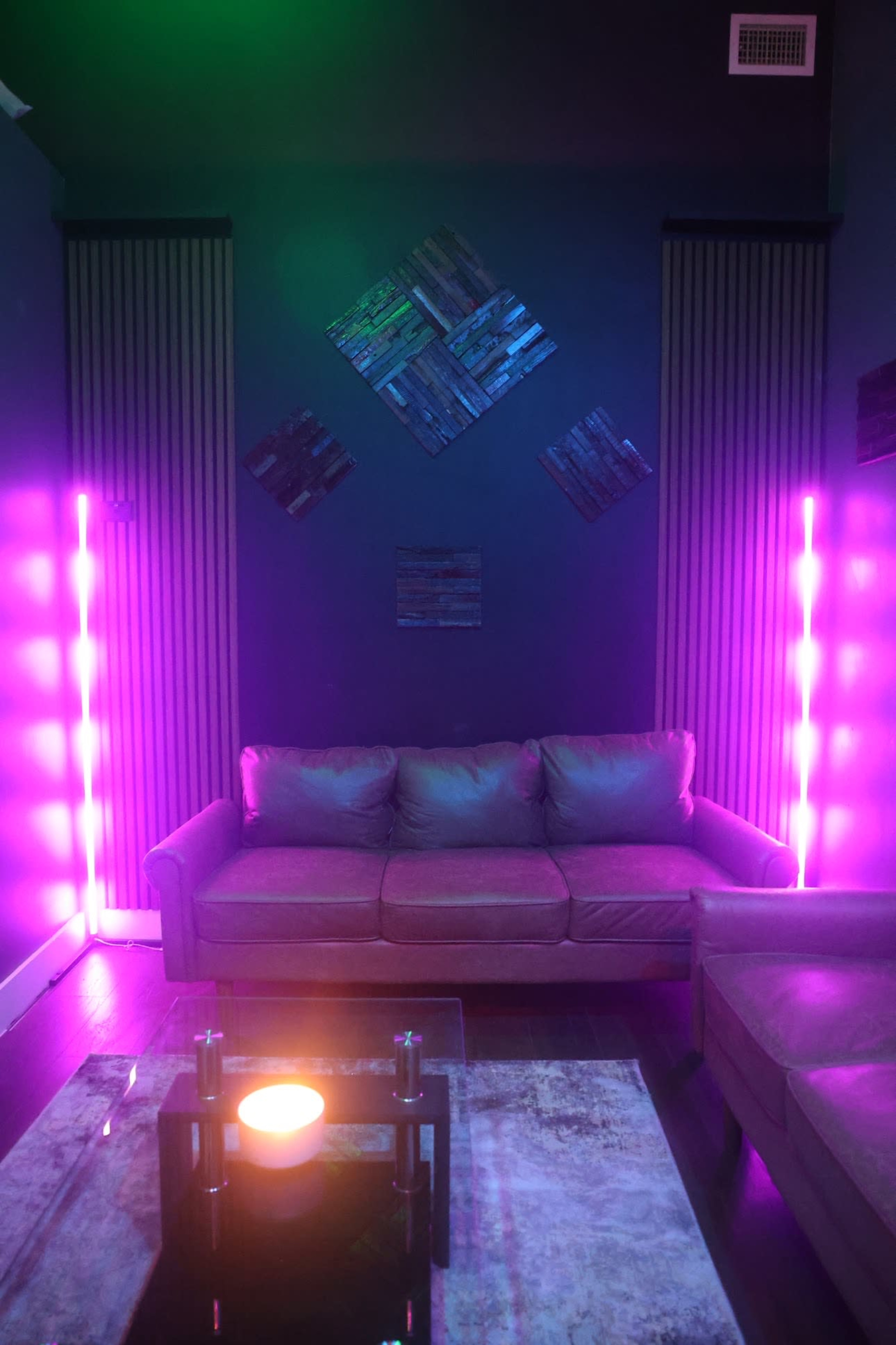A dimly lit room featuring a brown couch, a glass coffee table with a lit candle, and illuminated vertical light panels in purple hues against dark walls.