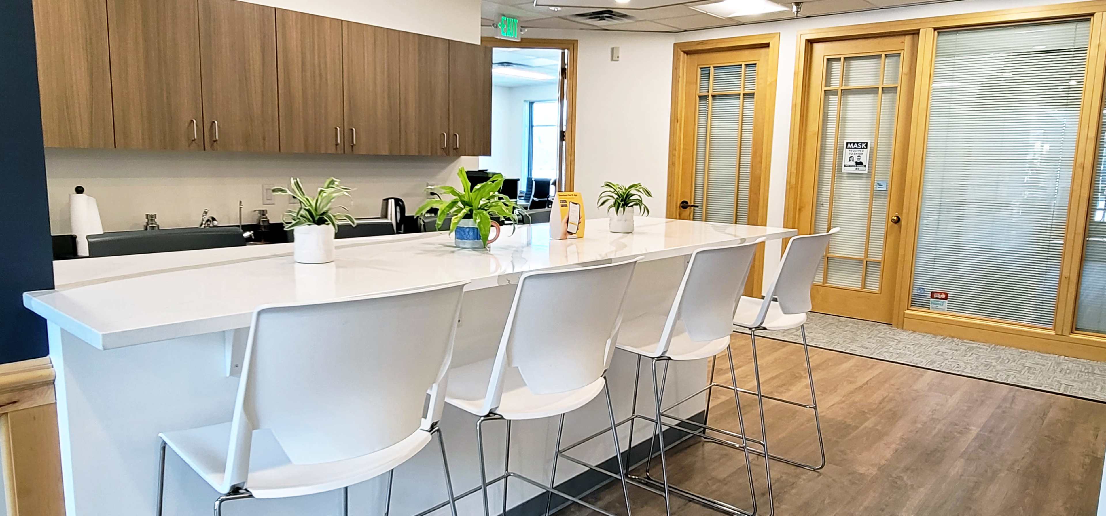 A modern office kitchen area with a white counter and several tall, white bar stools, featuring plants and wooden cabinetry.