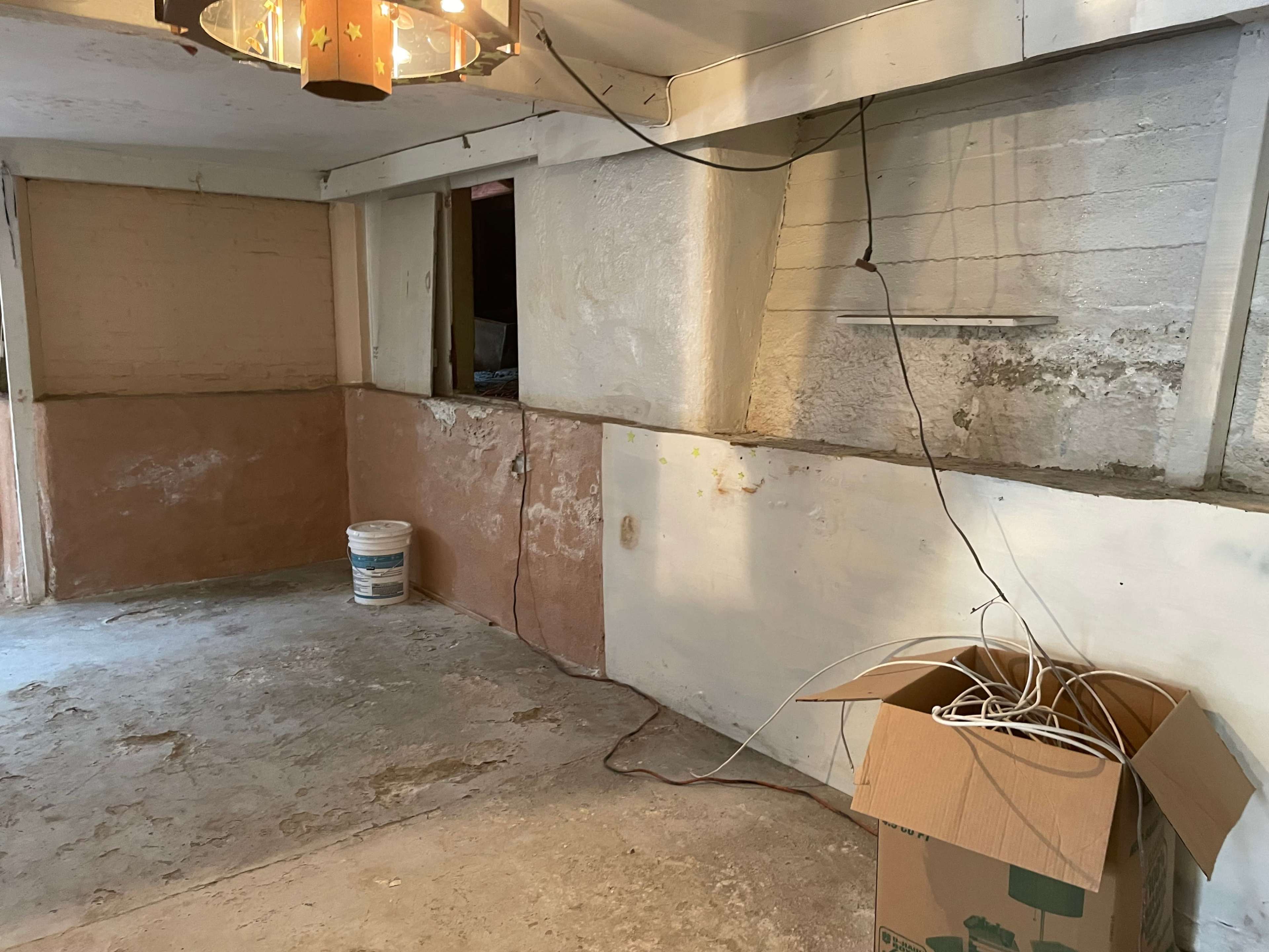 The image shows an unfinished room with bare walls, a bucket of paint, and a cardboard box containing cables.