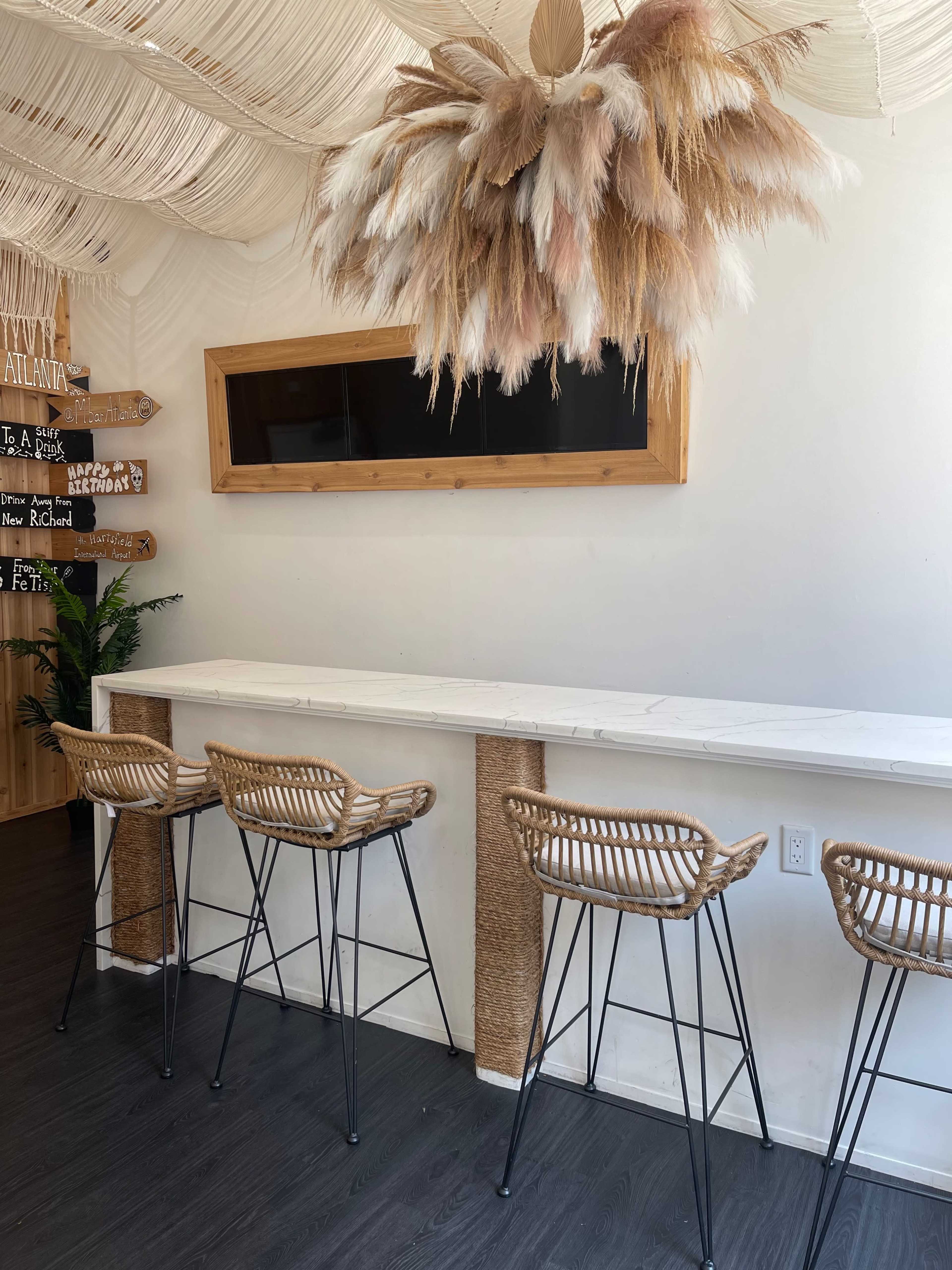 A minimalist bar area features four rattan stools lined up against a white marble counter beneath a large decorative ceiling fixture made of dried plants.