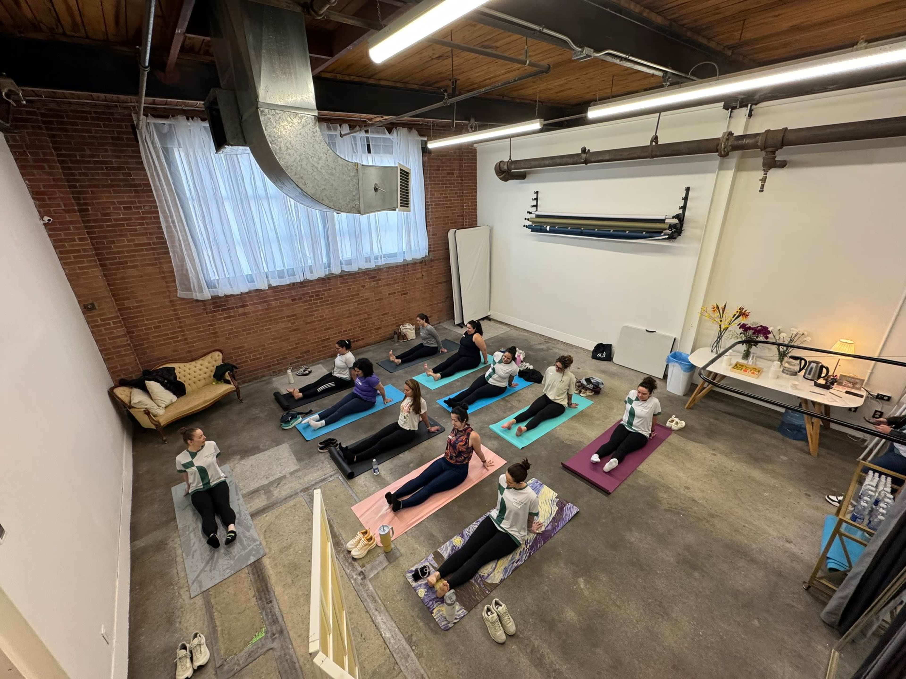A group of people is participating in a yoga class on mats in a spacious, well-lit studio with exposed brick walls.