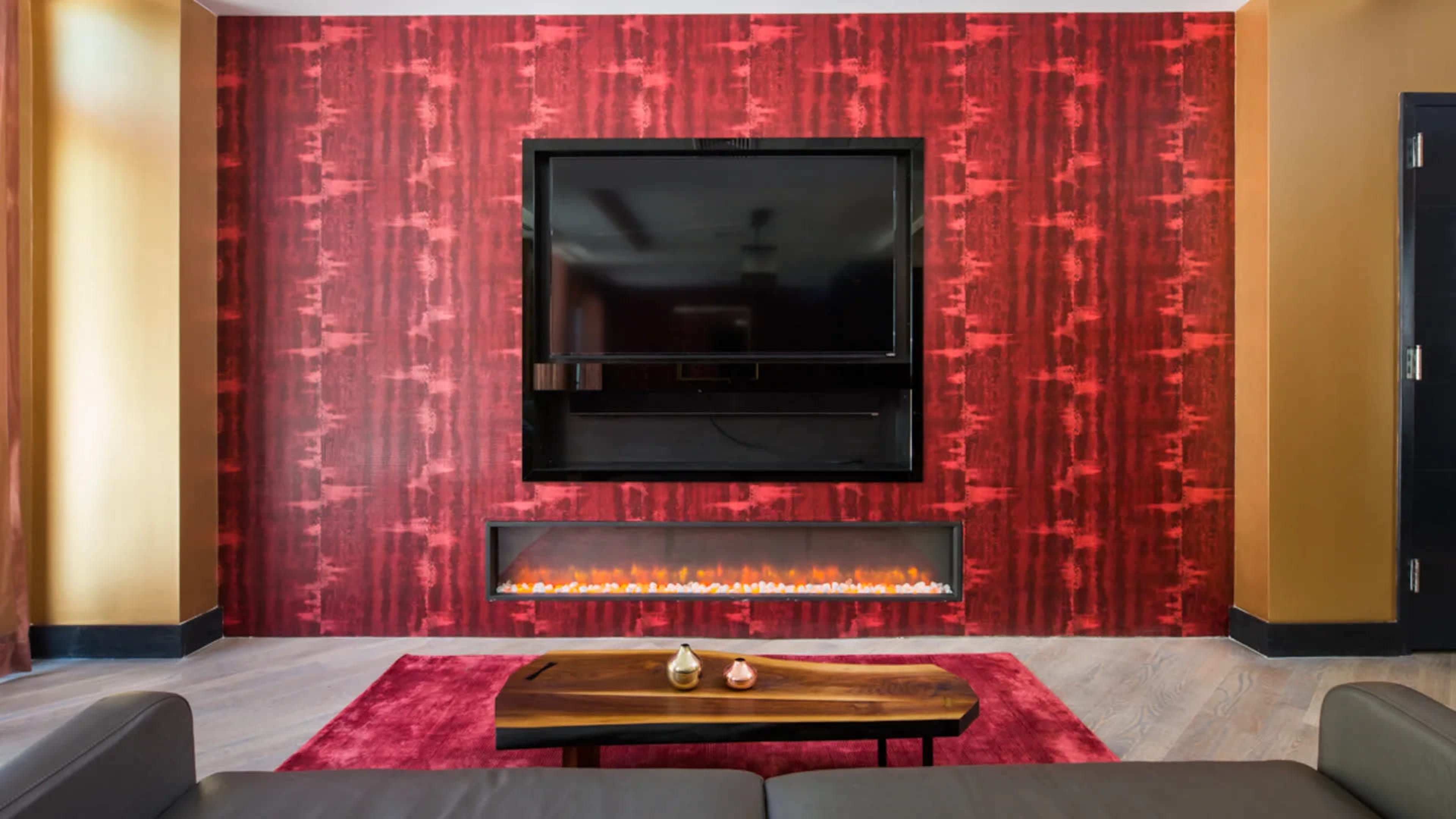 A living room with a red patterned wall, a mounted television, an electric fireplace below, and a coffee table in front.
