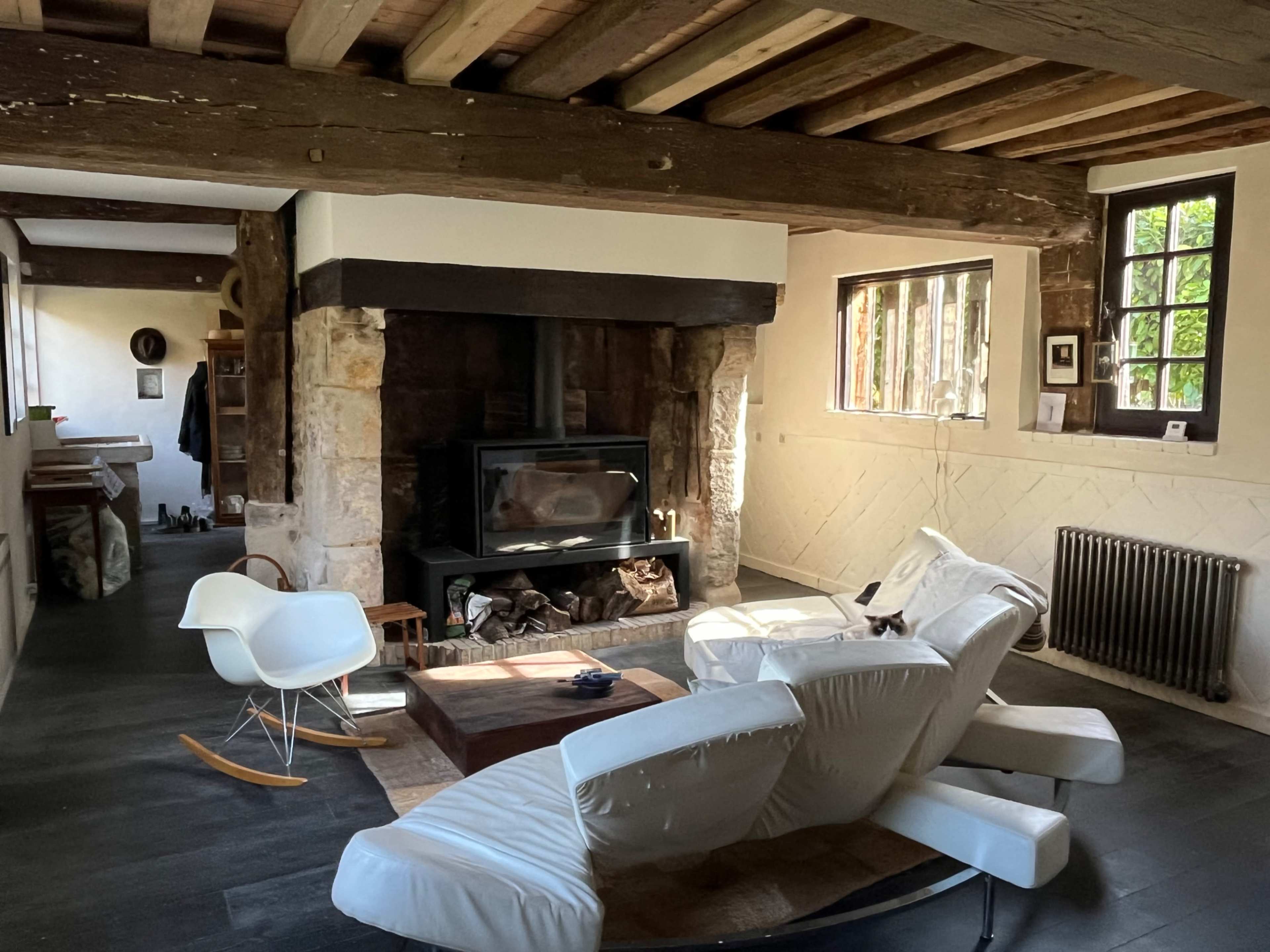 A modern living room features a white rocking chair, a wooden coffee table, and a stone fireplace surrounded by exposed wooden beams.
