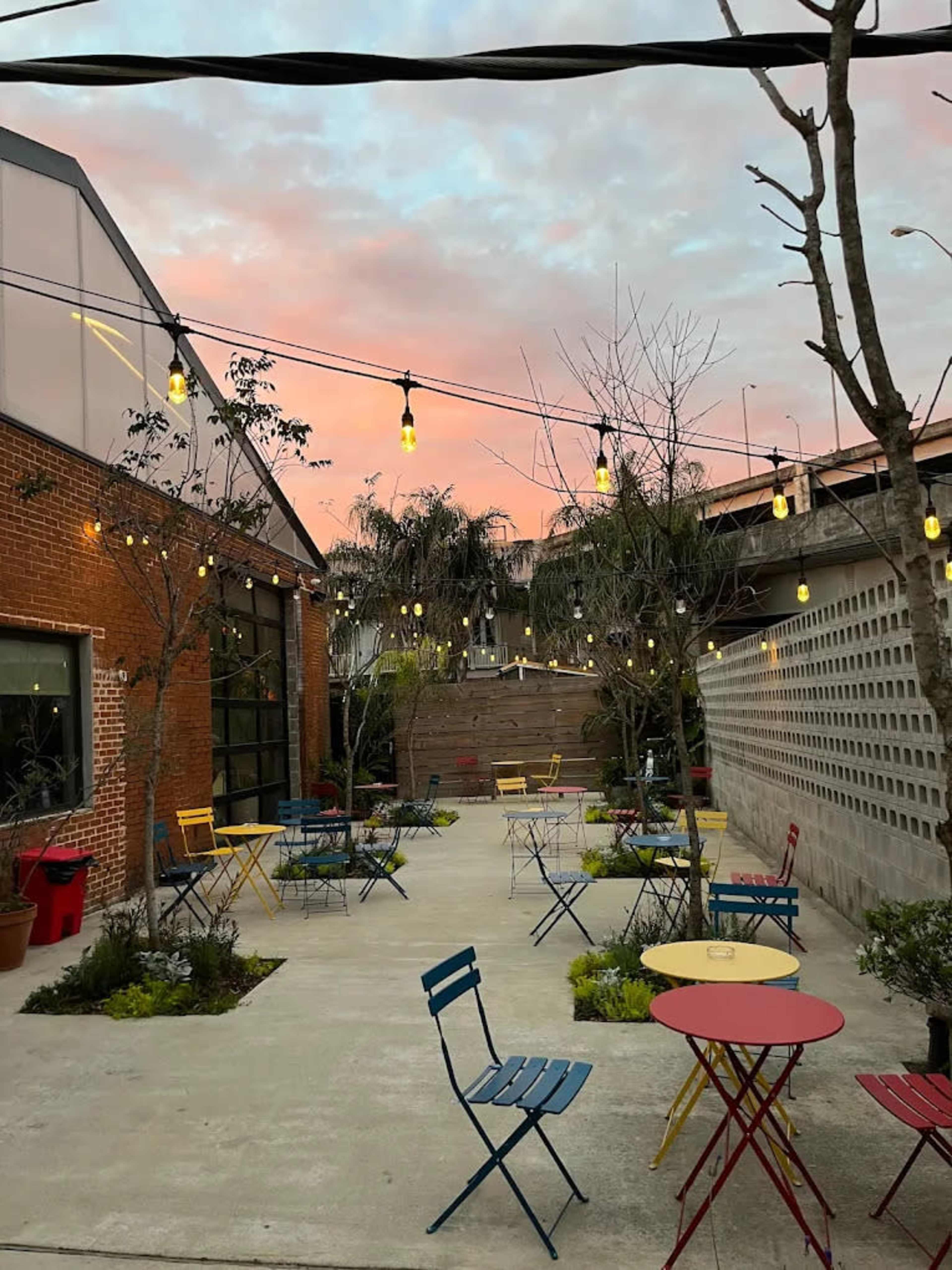 The image shows a small outdoor courtyard with colorful metal tables and chairs, surrounded by trees and string lights against a colorful sky at sunset.