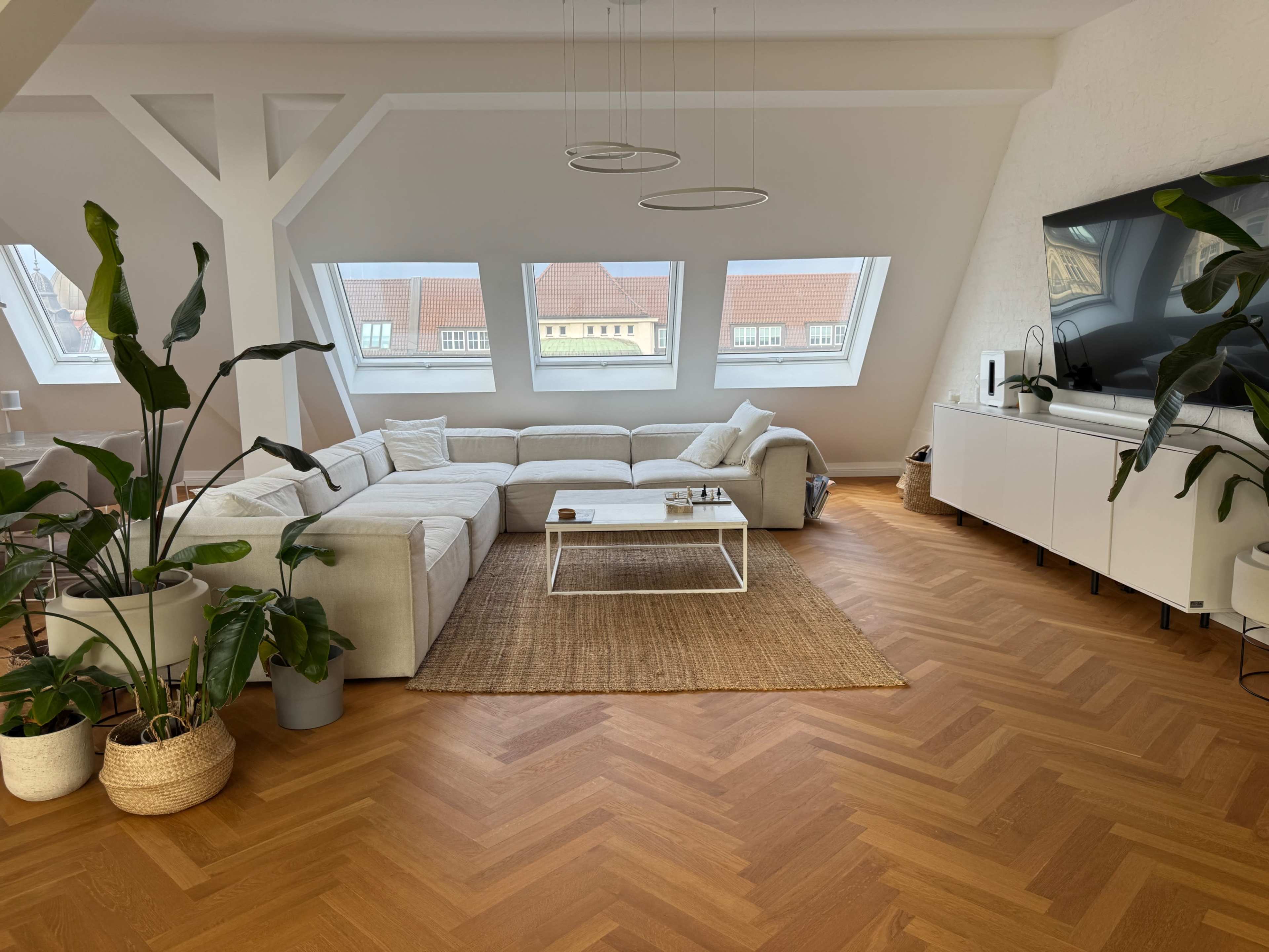 The image shows a modern living room with a sectional sofa, a coffee table, and large windows, complemented by plants and a television unit against the wall.