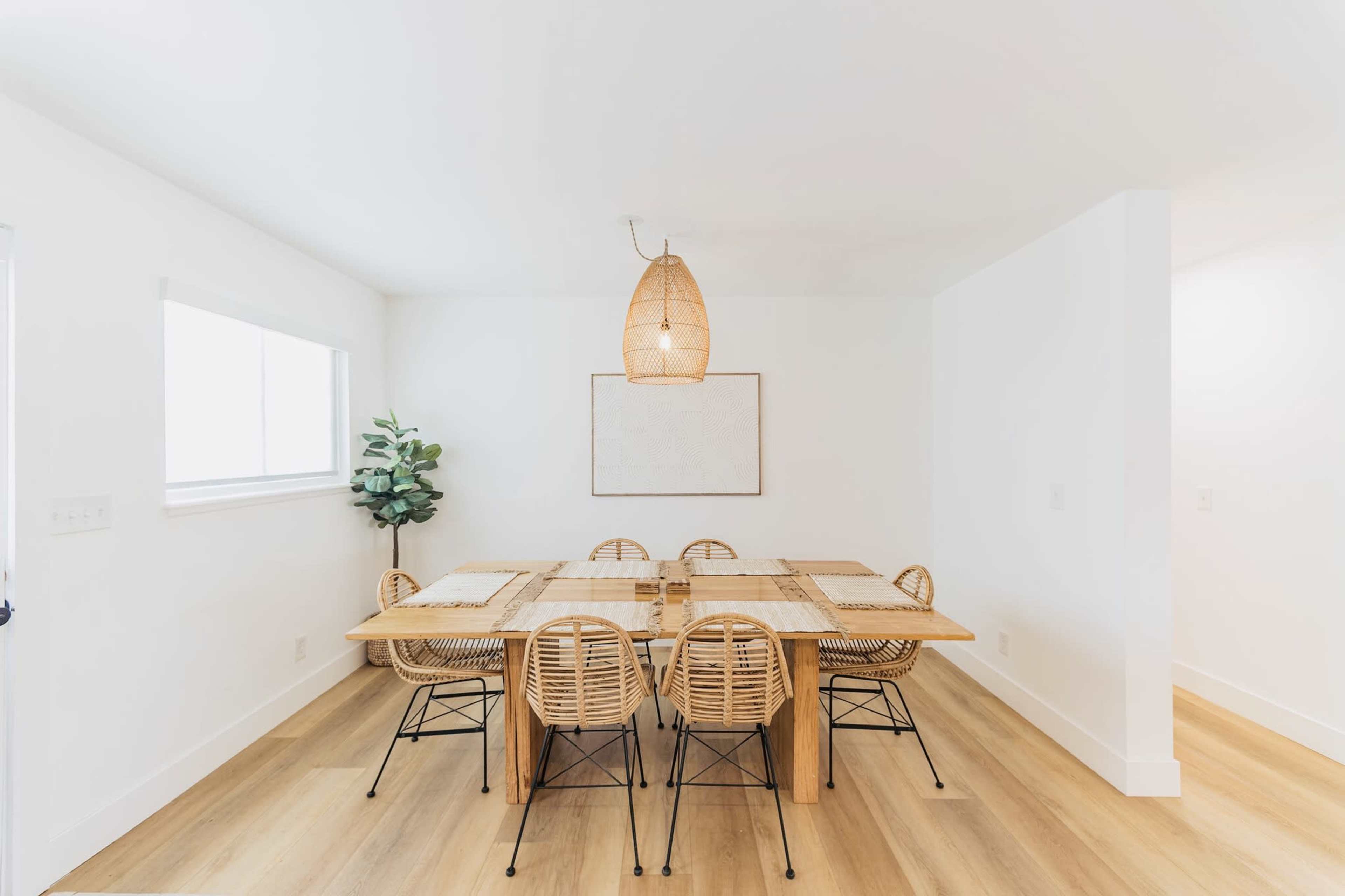 A dining room features a wooden table surrounded by six rattan chairs, with a pendant light above and a potted plant in one corner.