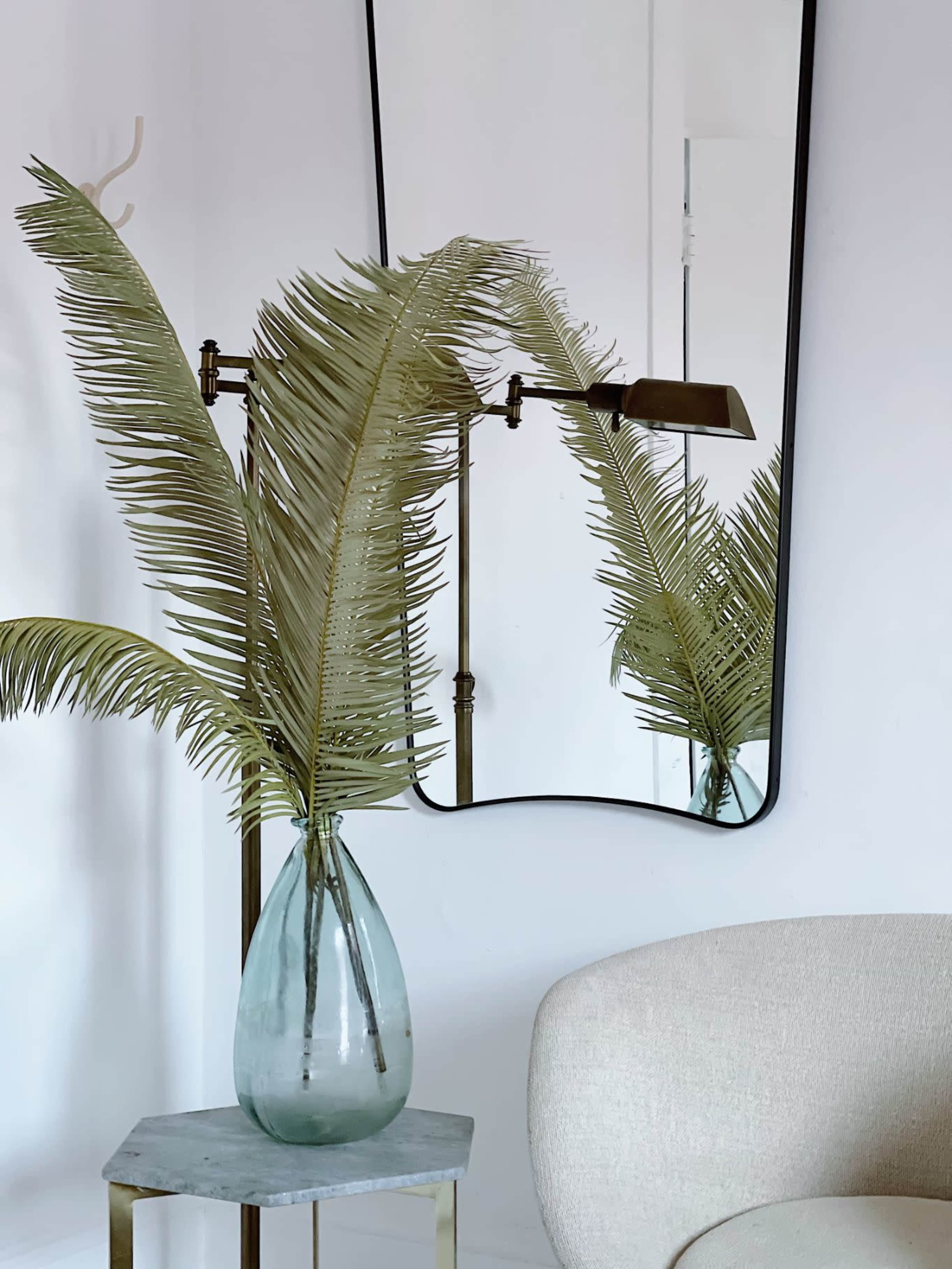 A tall vase with palm fronds is positioned on a small table next to a minimalist chair, with a wall mirror reflecting the scene.