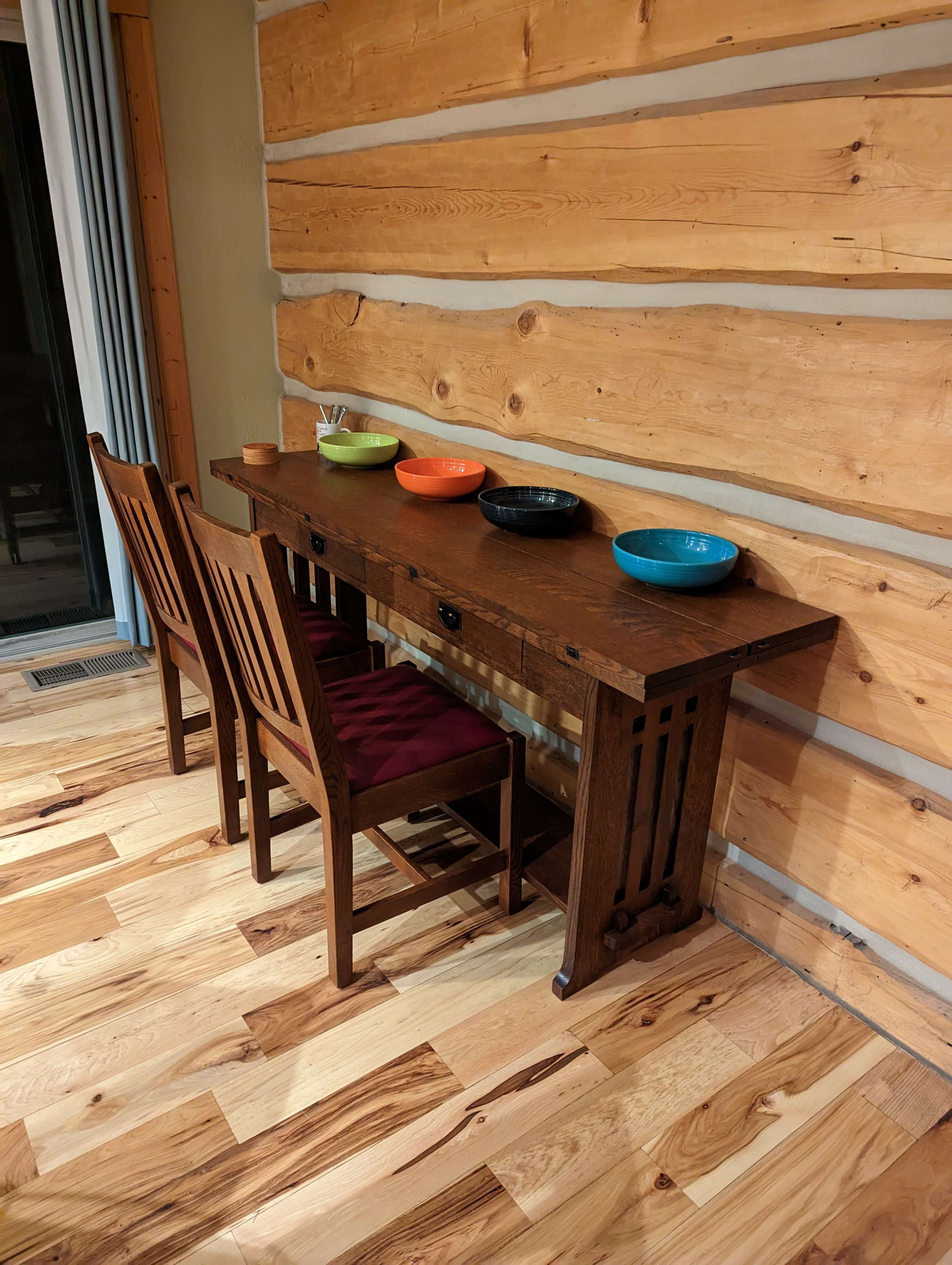 A wooden table with two chairs is placed against a log cabin wall, featuring four colorful bowls arranged on top.
