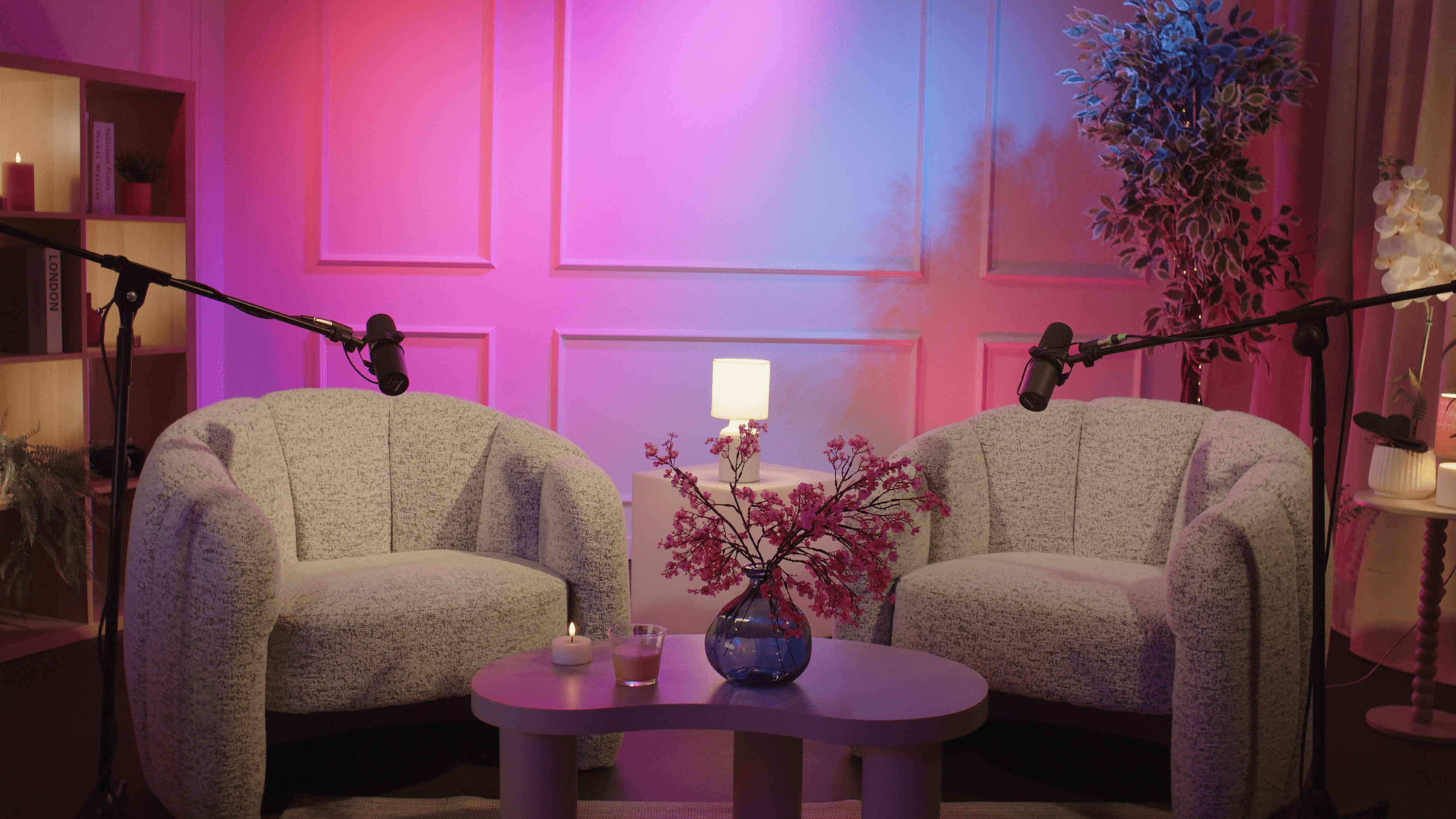 A brightly lit interview setup features two cozy armchairs with microphones attached, a small table in between them holding a vase of flowers, and soft ambient lighting in pink and purple tones.