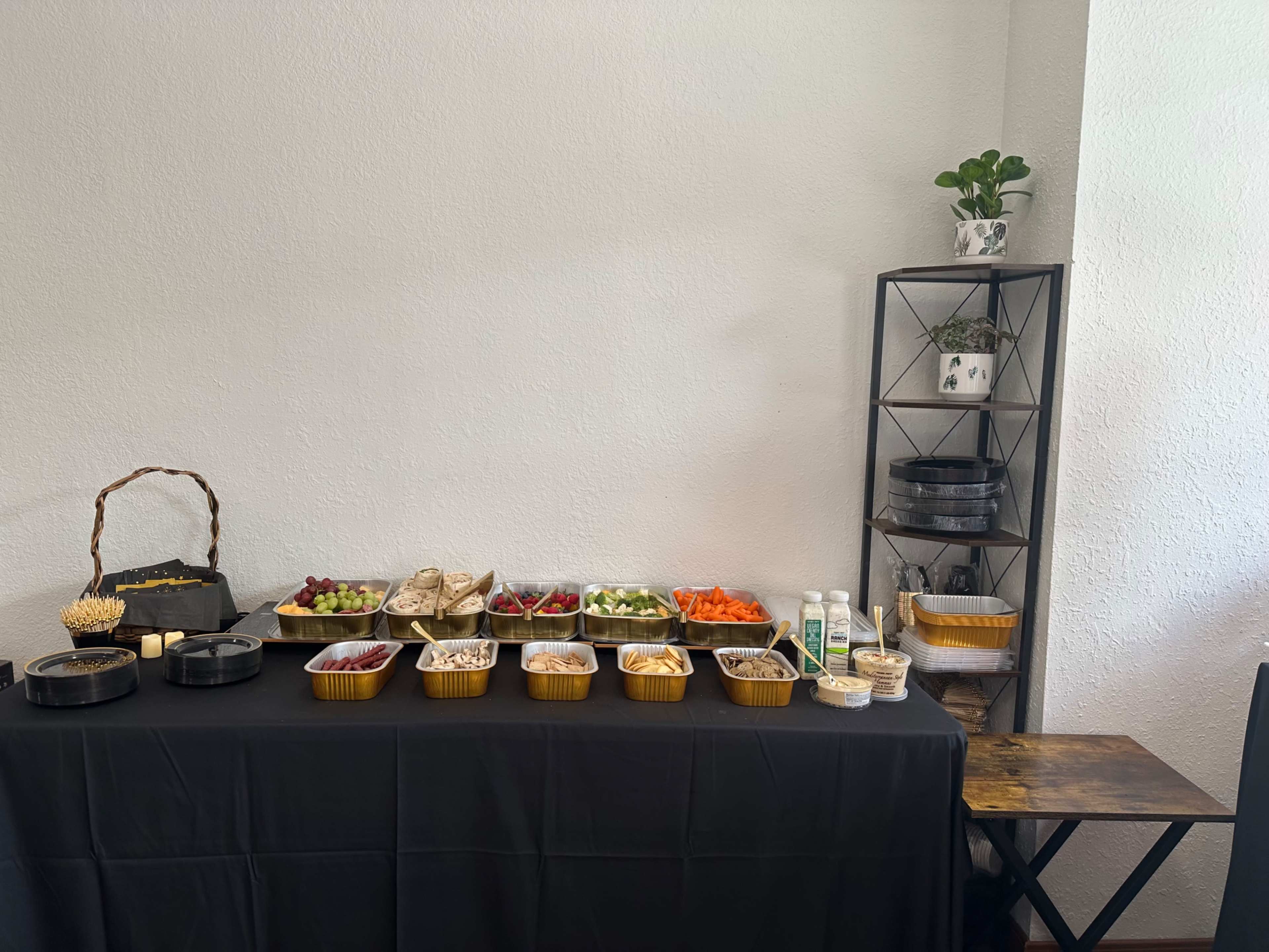 A table covered with a black cloth displays various snacks and fruits in containers, alongside a small shelf with potted plants.