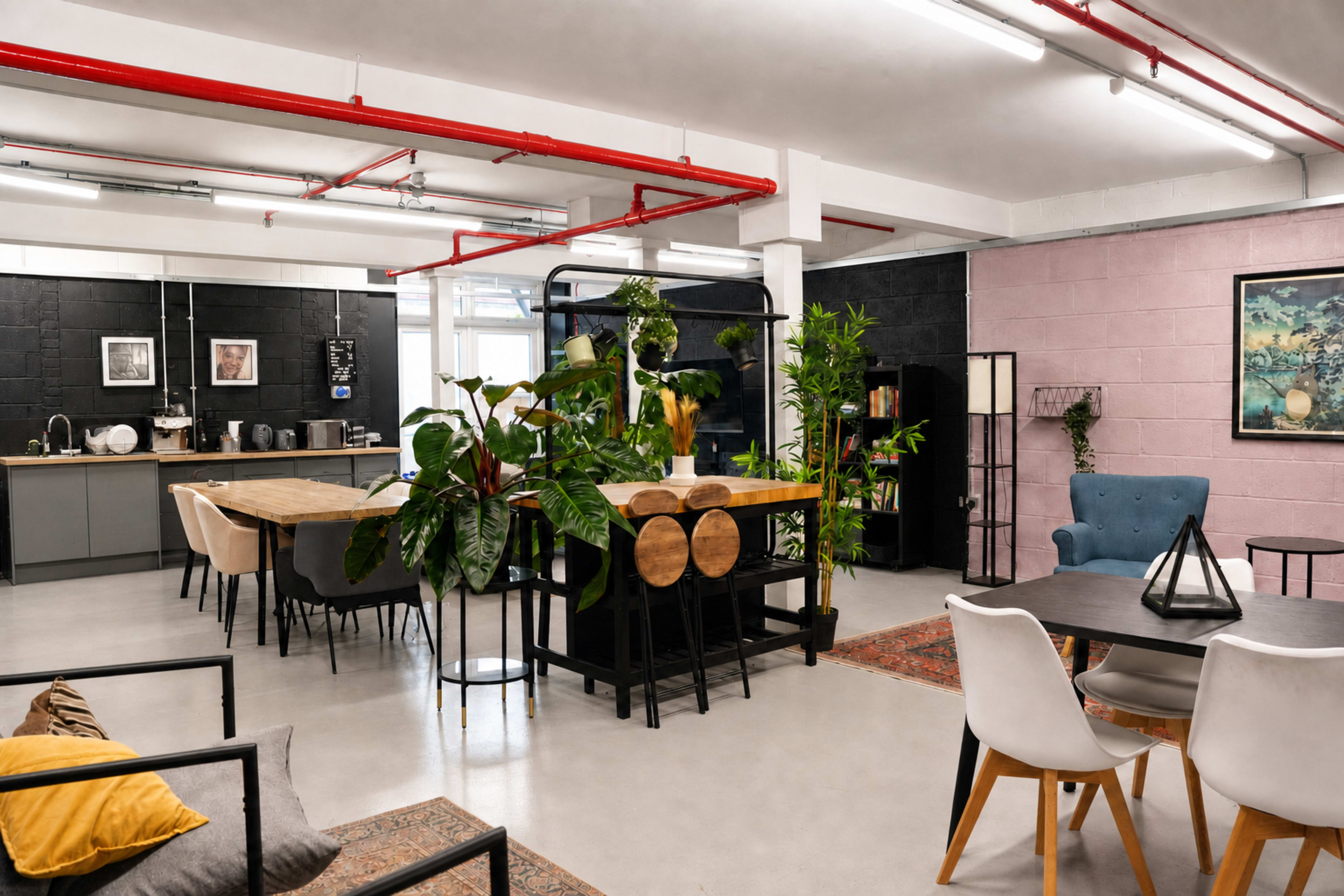 The image shows a modern, open-plan office space with a kitchen area, several tables, and various plants decorating the environment.