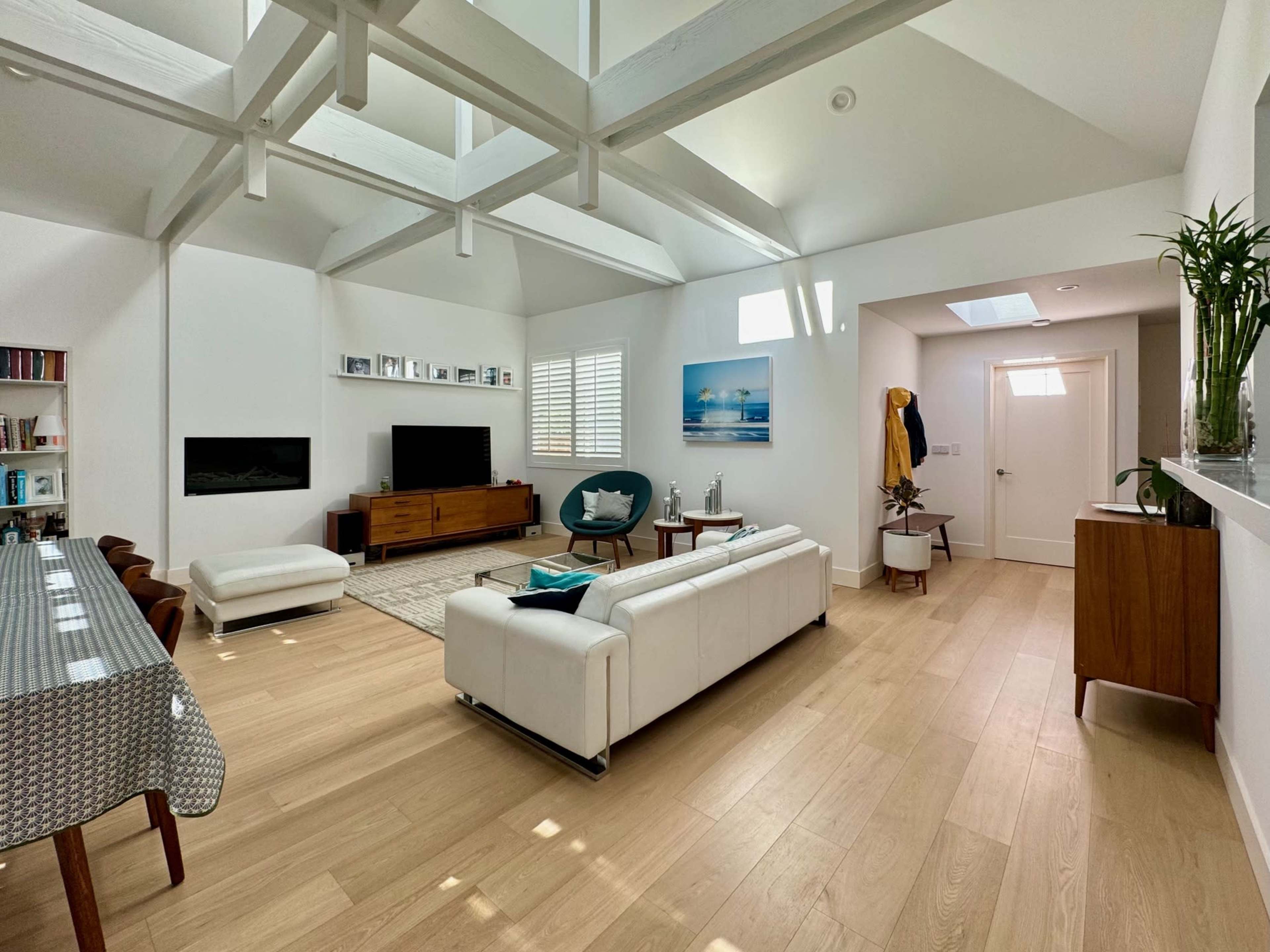 The image shows a bright, modern living room with a white couch, a wooden TV unit, a dining table, and large windows allowing natural light to fill the space.