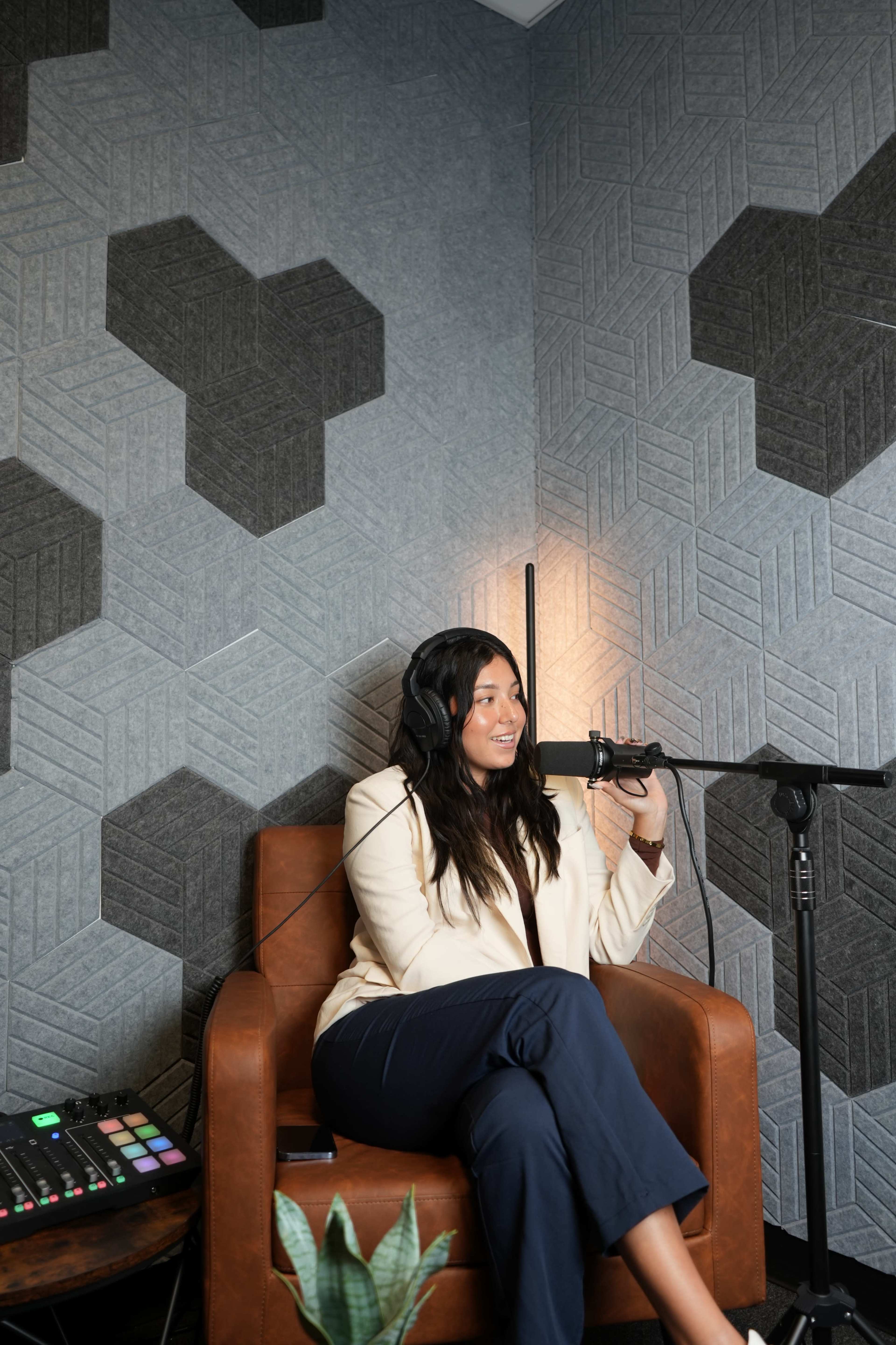 A woman sits in a brown leather chair, wearing headphones and speaking into a microphone, with geometric patterned wallpaper in the background.